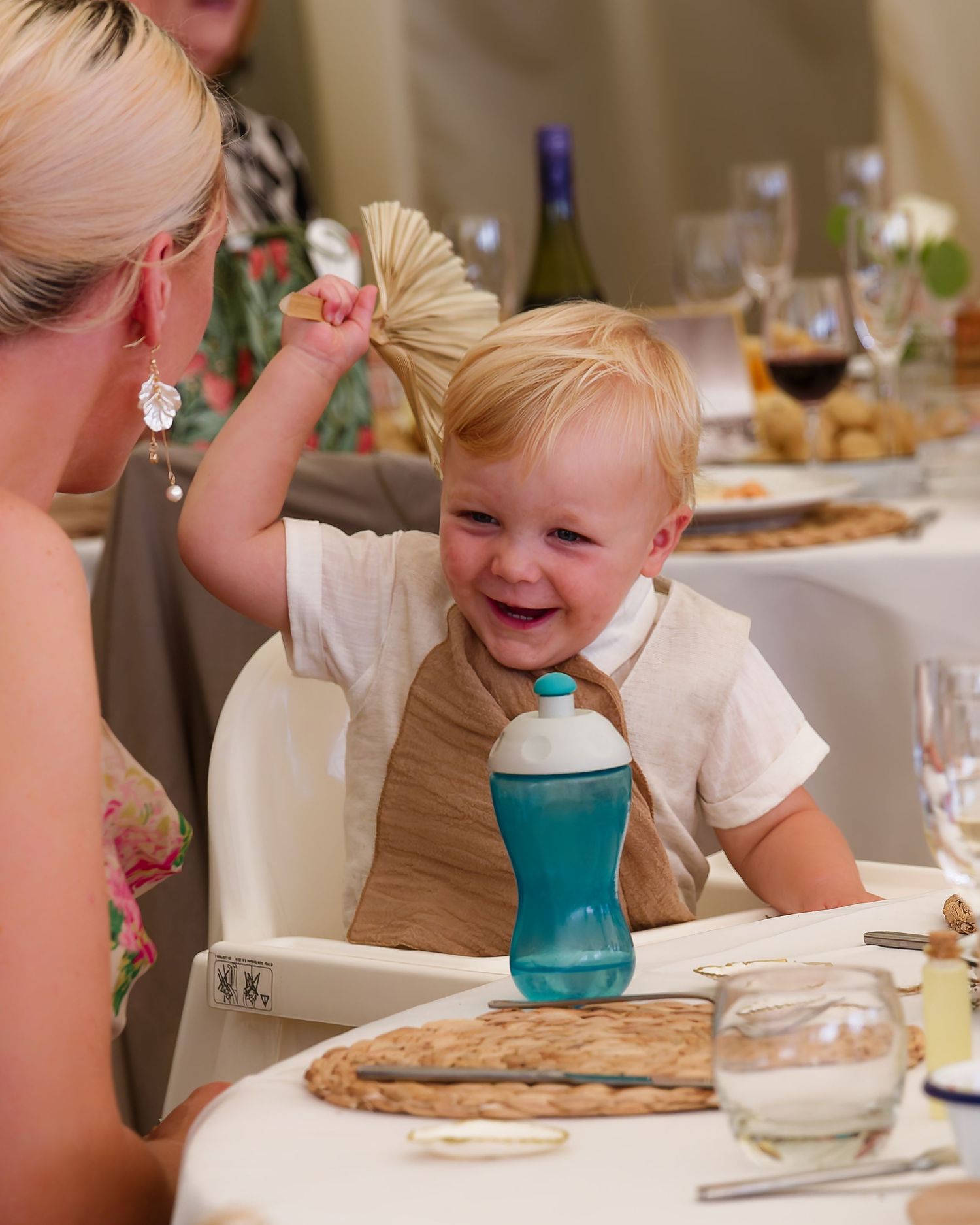 Laughing baby in high chair waves a palm leaf fan at a wedding reception dinner table.