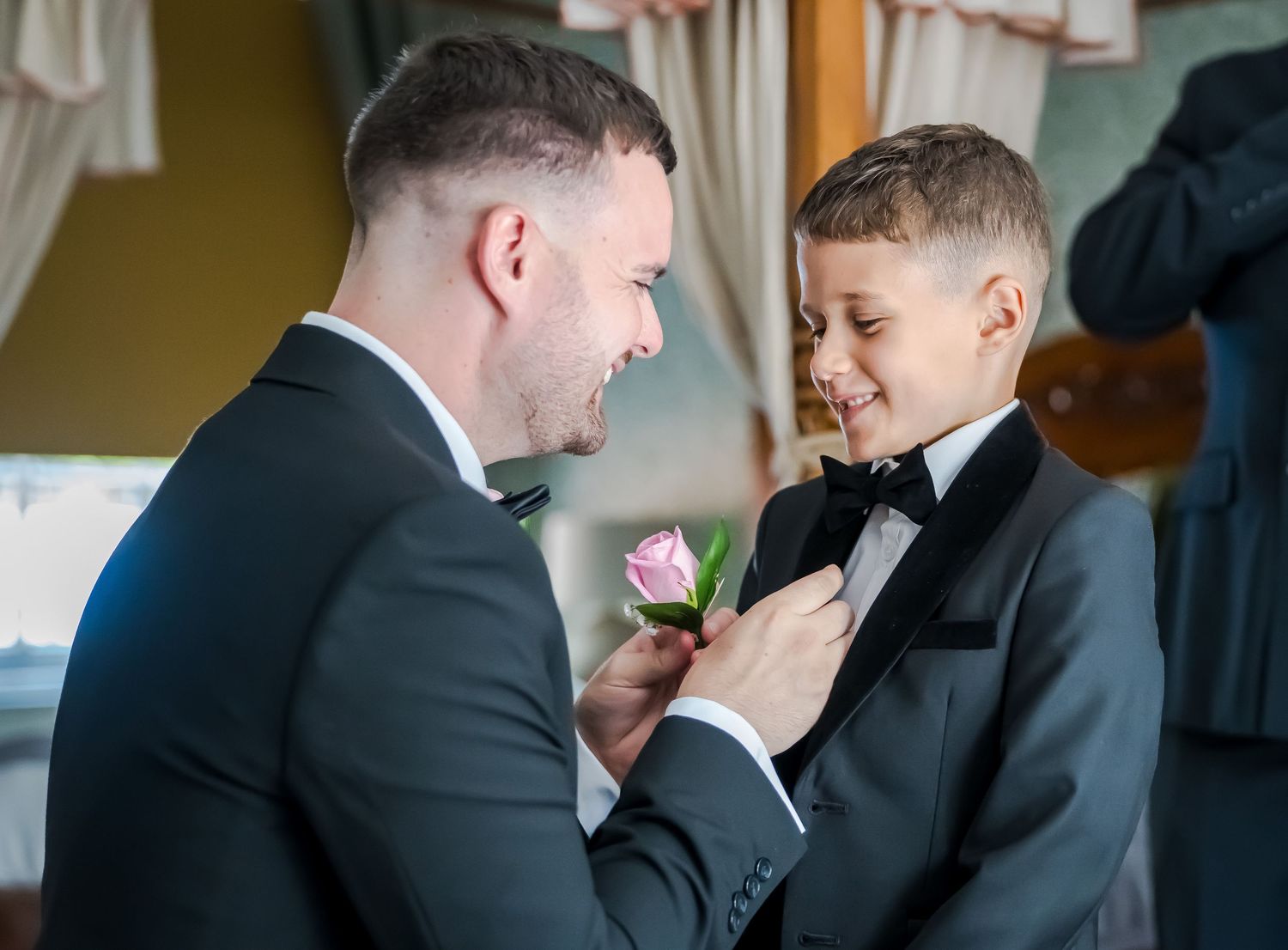 Father pinning a pink boutonniere on a young boy in a tuxedo at a wedding ceremony.