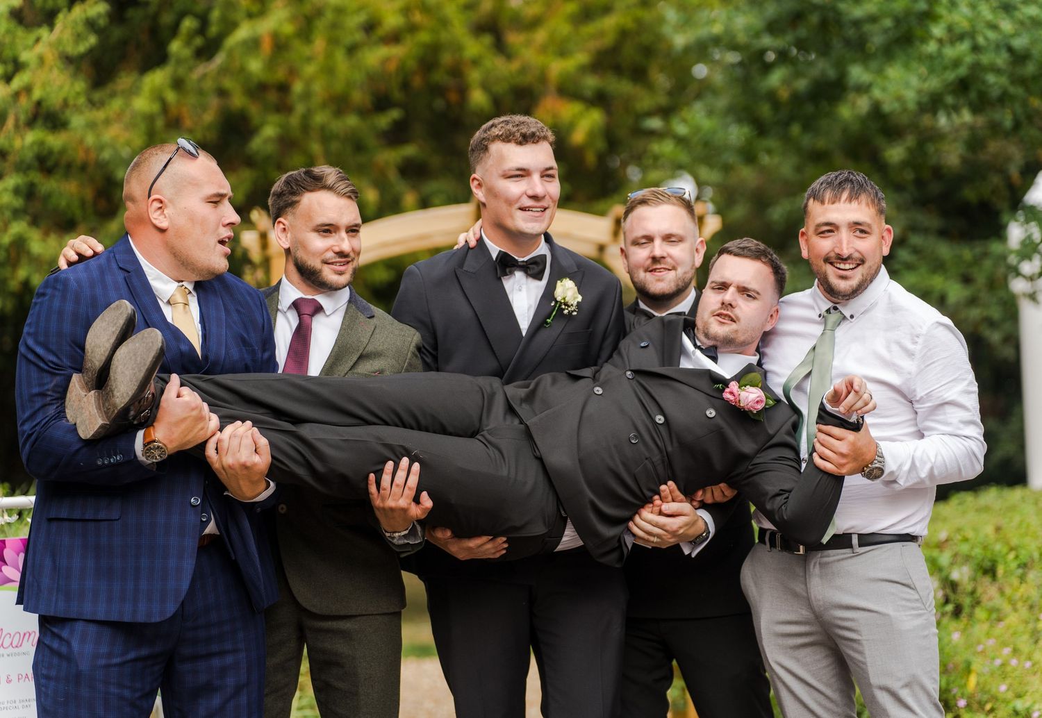 Groomsmen laughing while carrying groom in suit at outdoor wedding celebration.