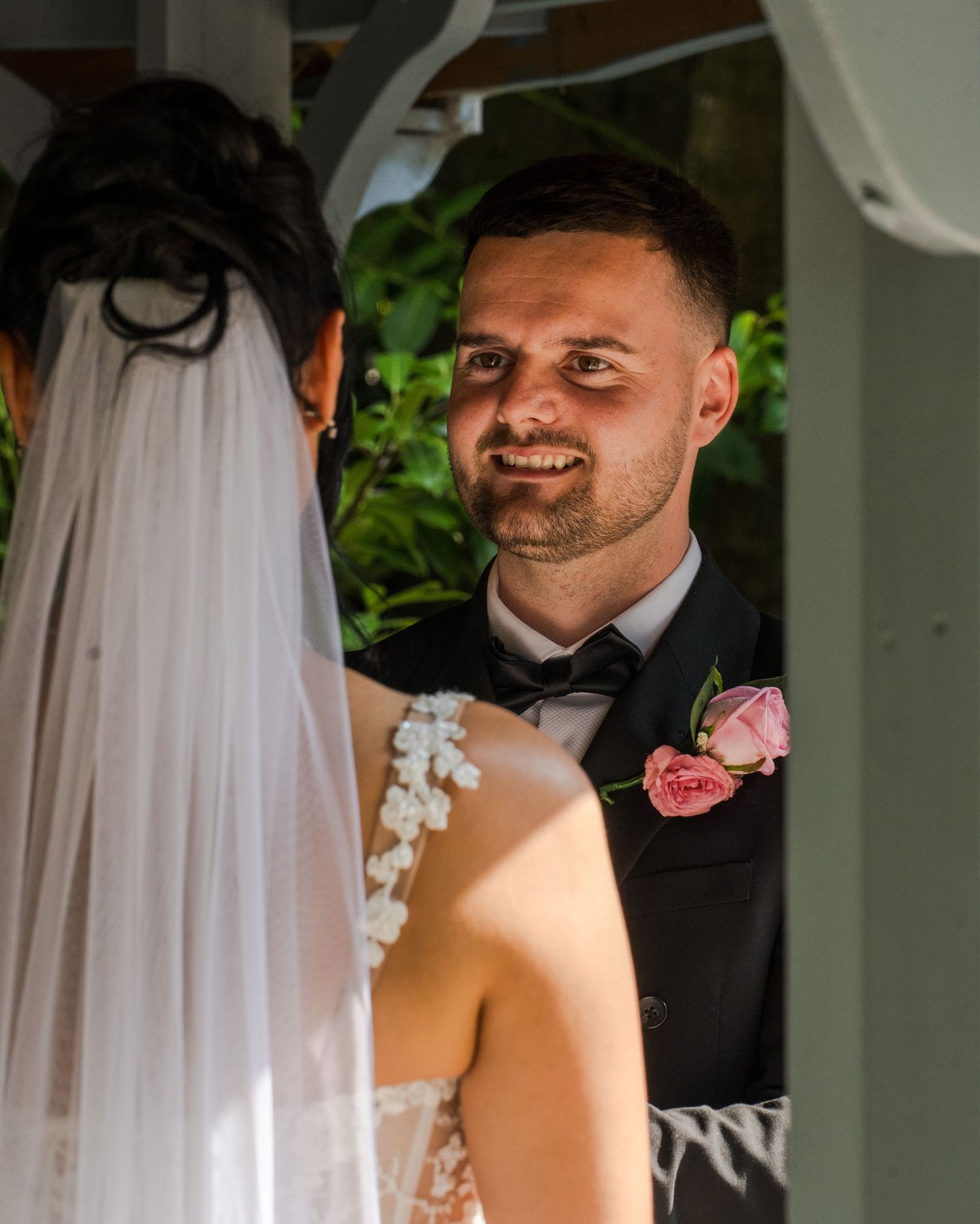 Groom in black tuxedo with pink rose boutonniere smiling at bride in white lace dress and veil during wedding ceremony.