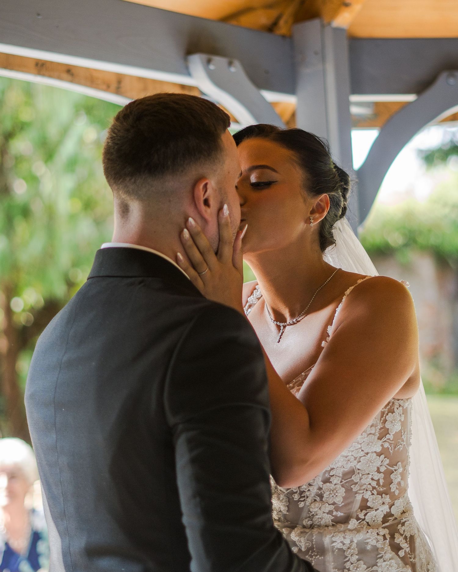 Bride and groom share their first kiss during an outdoor wedding ceremony under a wooden pergola.