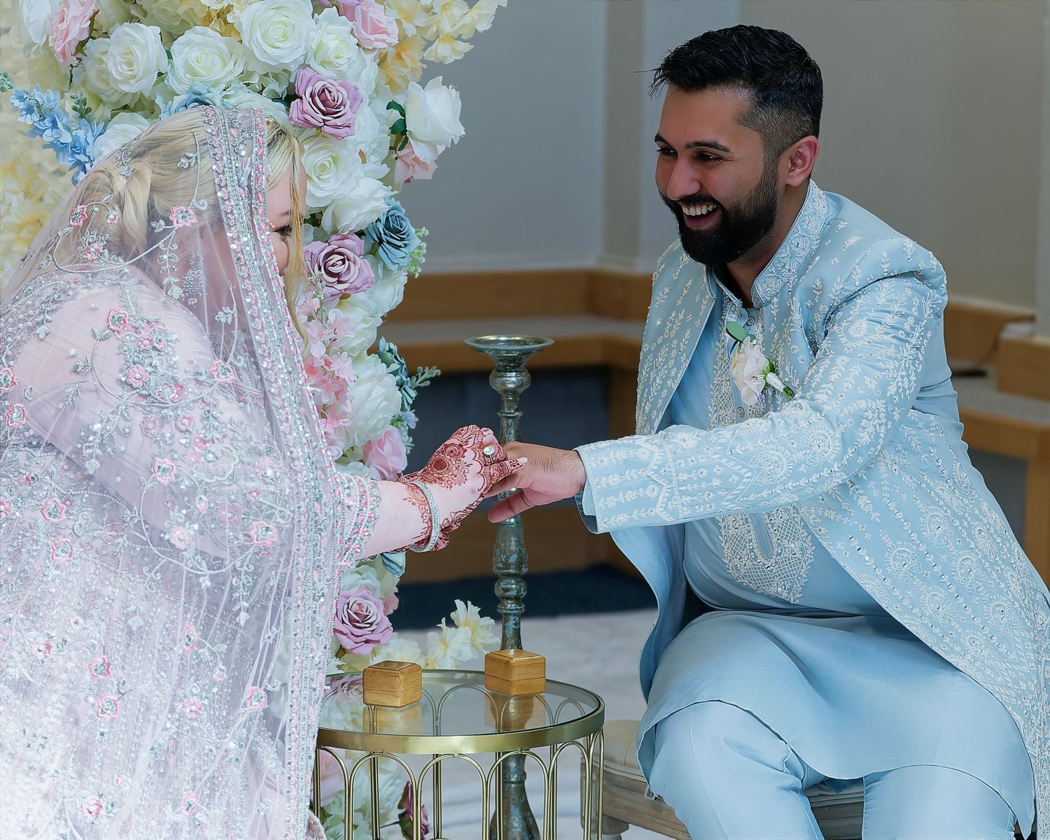 A groom in a light blue suit exchanges rings with a bride in a white hijab and embroidered gown at a floral wedding ceremony.