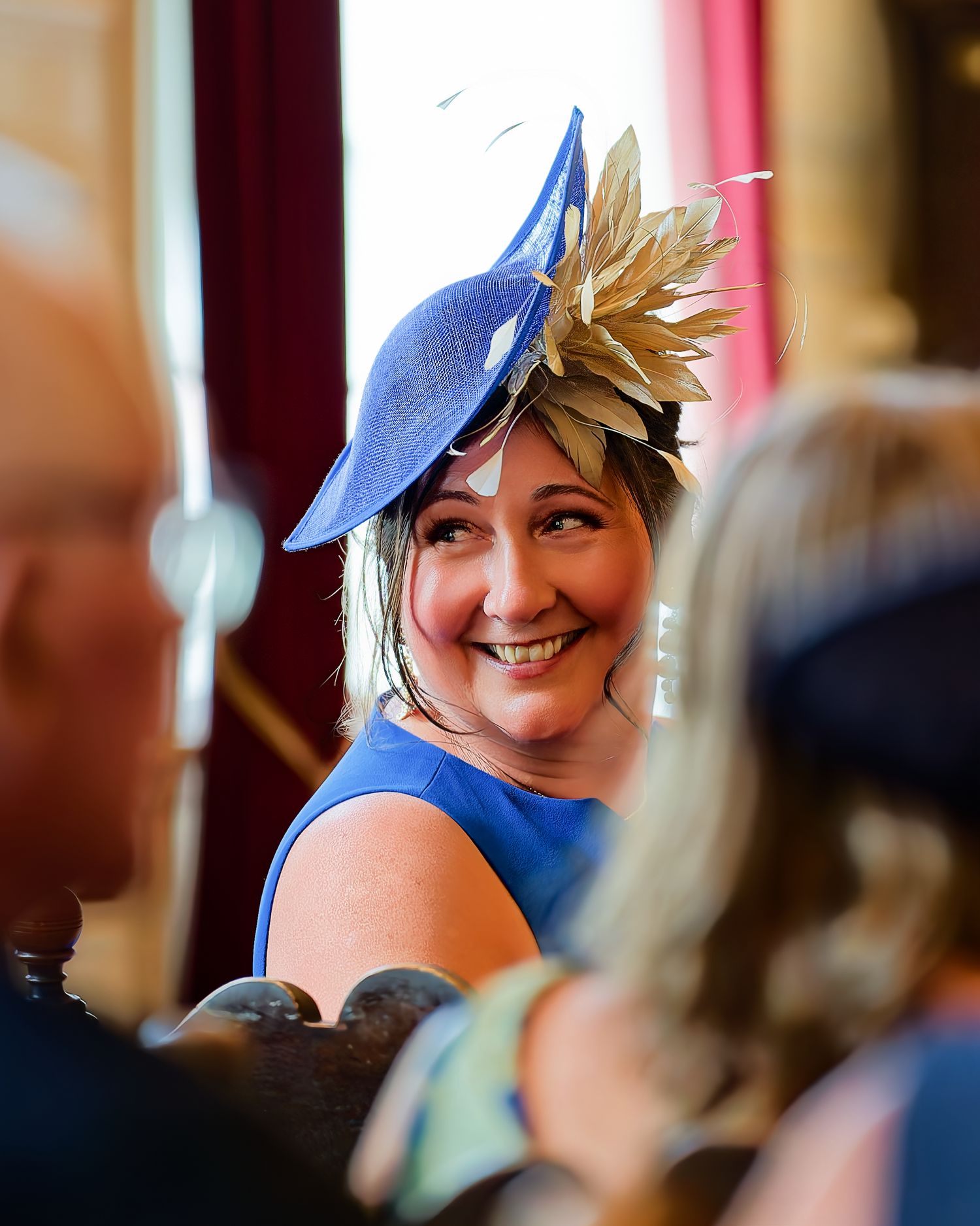 Smiling woman wearing a blue fascinator hat with gold leaves at a wedding reception.