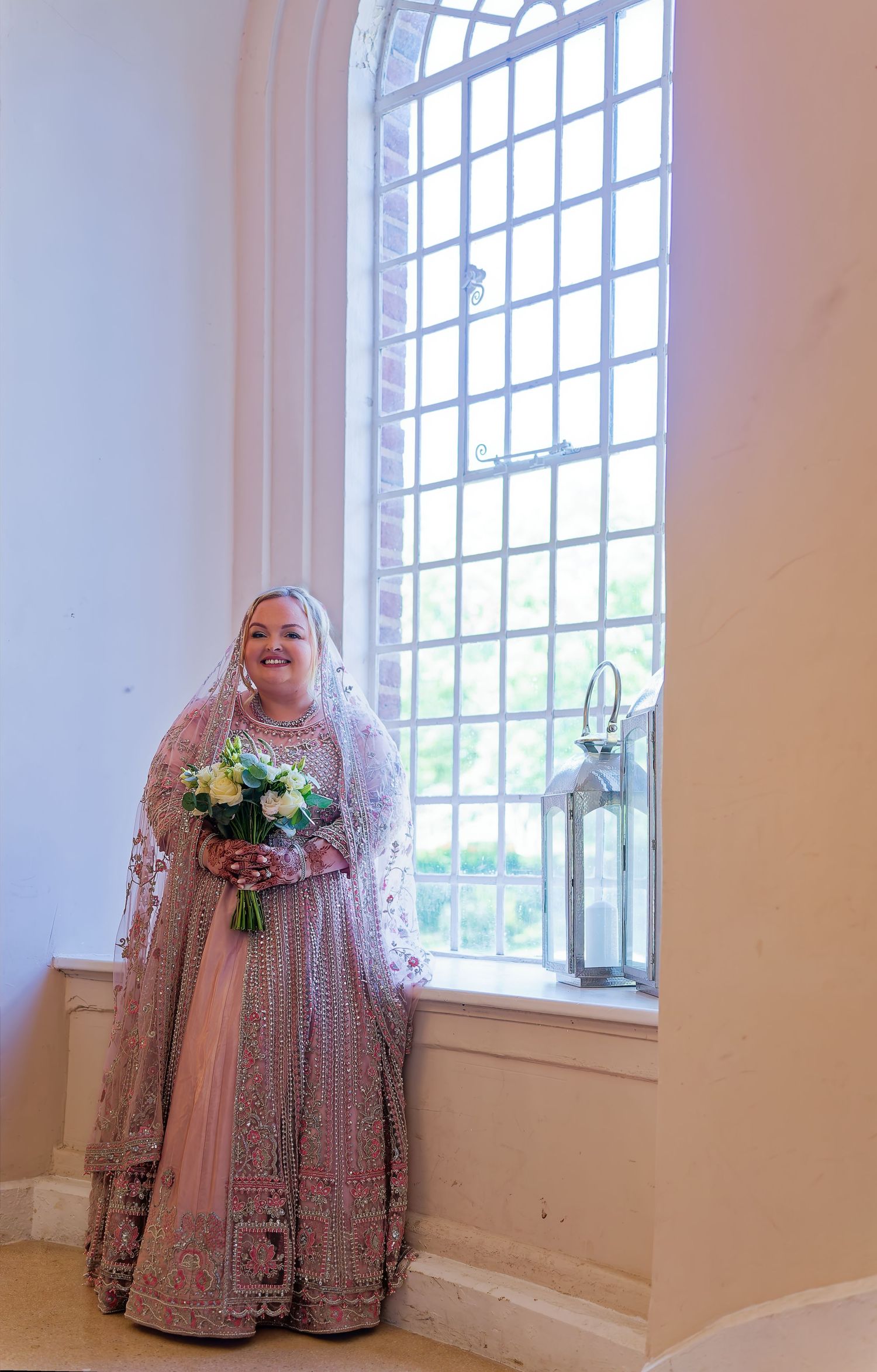 Smiling bride in pink embroidered lehenga holding bouquet by arched church window.