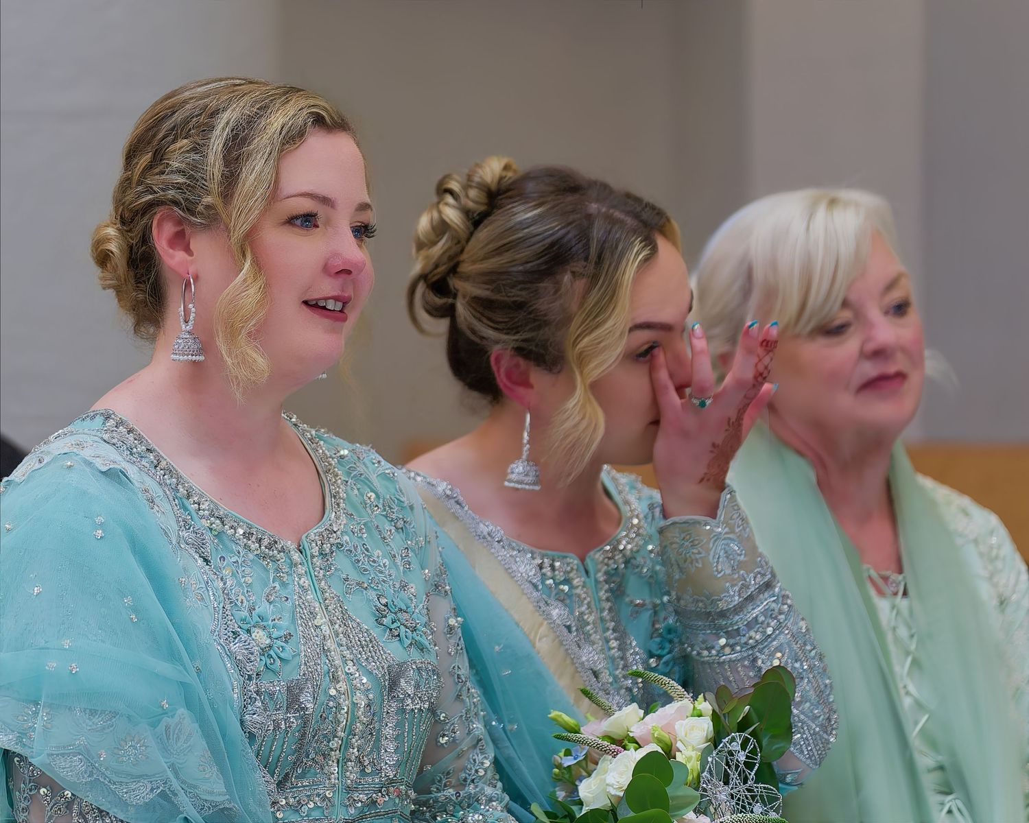 Three women in matching teal gowns at a wedding ceremony, one wiping tears, showing emotional joy.