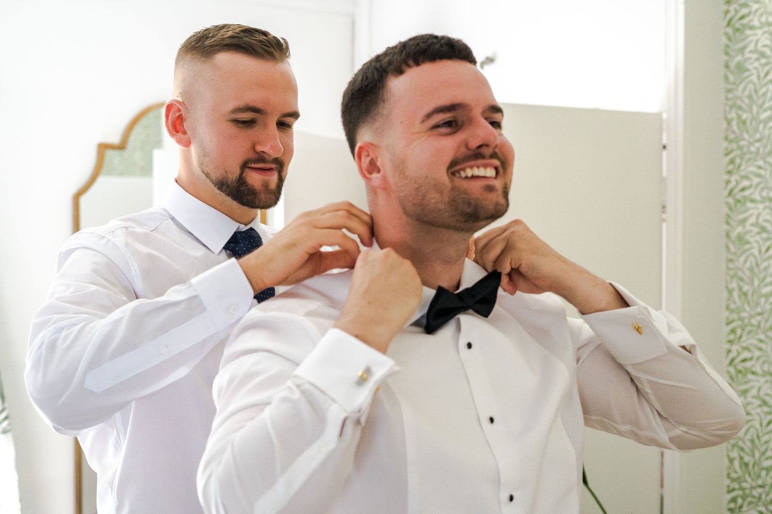 Two groomsmen in white shirts adjust a black bow tie before a wedding ceremony.