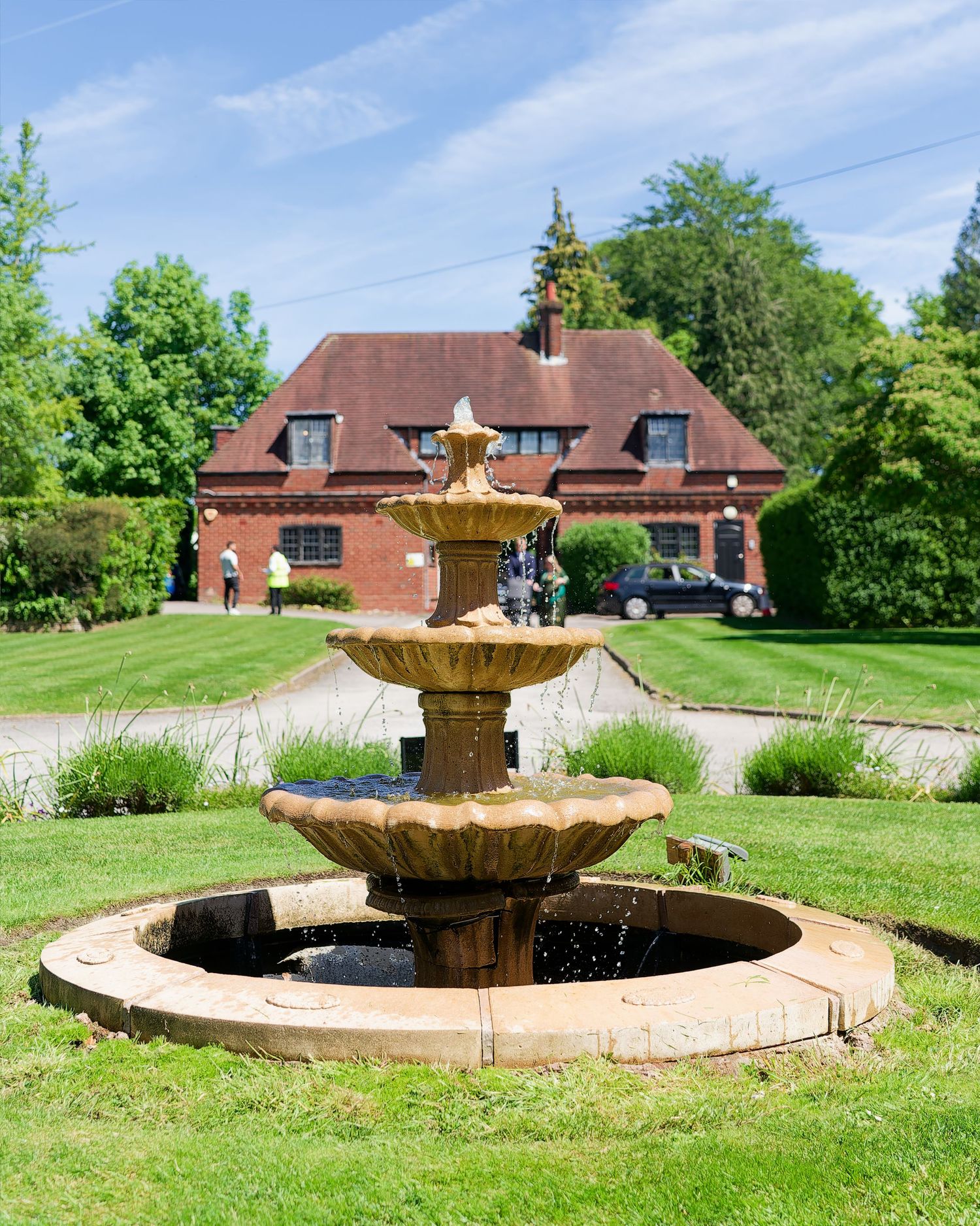 A three-tiered stone fountain on a green lawn with a red-brick country house and trees in the background.