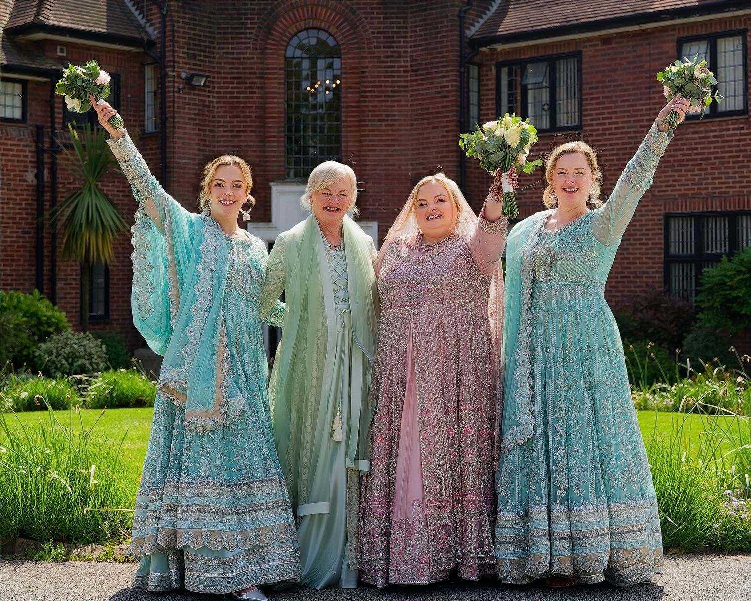 Four women in colorful Indian-style gowns celebrate outdoors, holding bouquets aloft, smiling joyfully near a brick building.