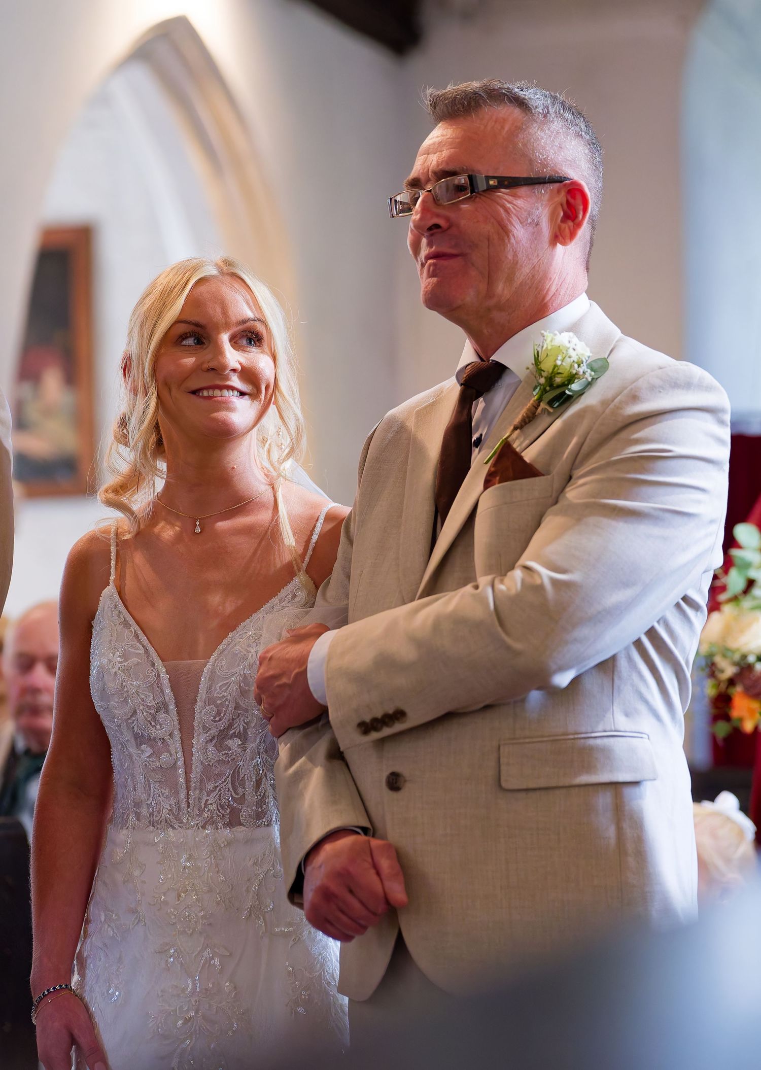A smiling bride in a white gown walks down the aisle with an older man in a beige suit at a church wedding.