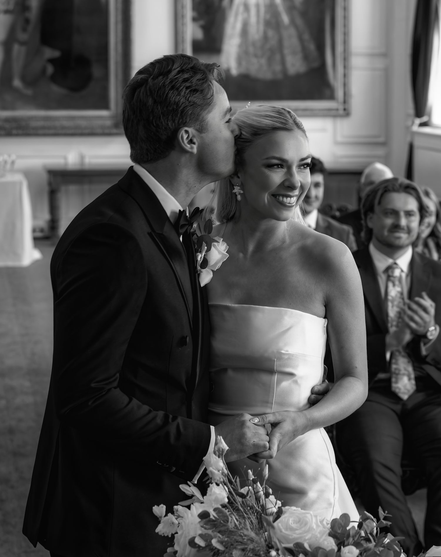 Black and white wedding photo of groom kissing bride on cheek as she smiles holding bouquet during ceremony.