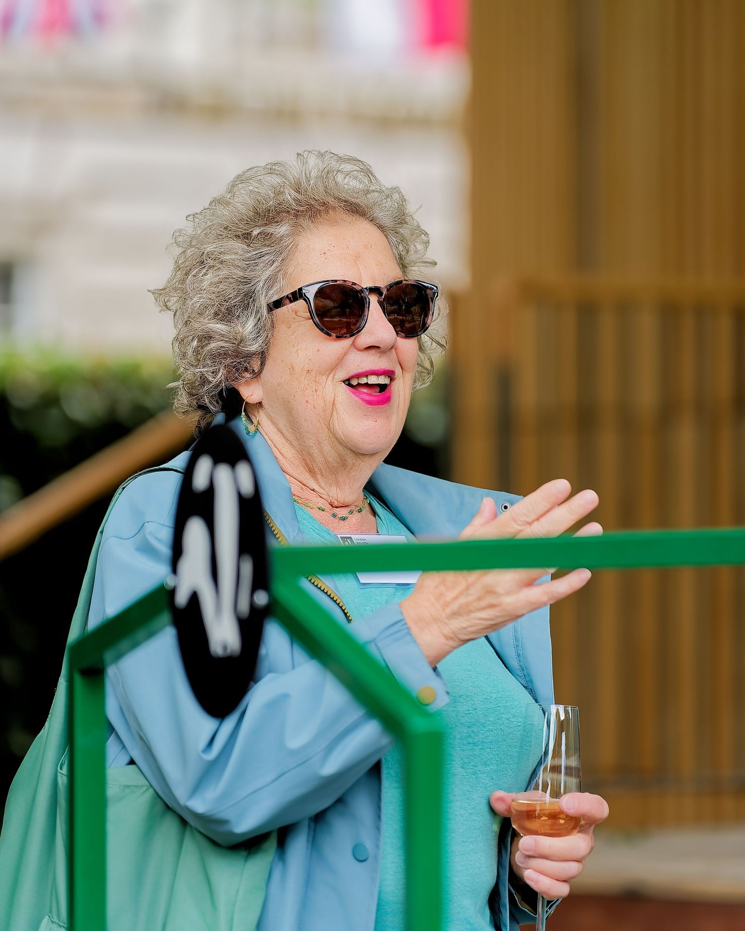 Smiling older woman with curly gray hair and sunglasses gestures while holding a glass of rosé wine outdoors in a light blue jacket.