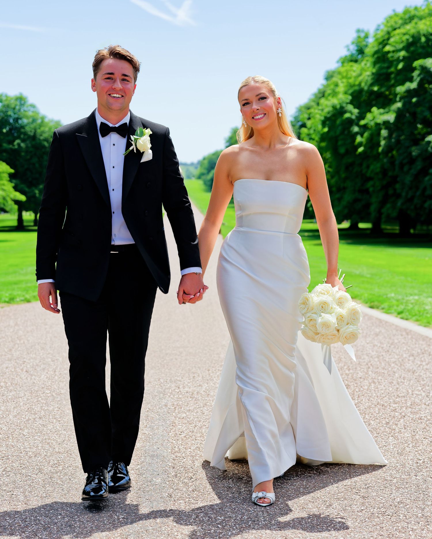 A smiling couple walks hand-in-hand on a sunny path, bride in strapless white gown holding bouquet, groom in black tuxedo.
