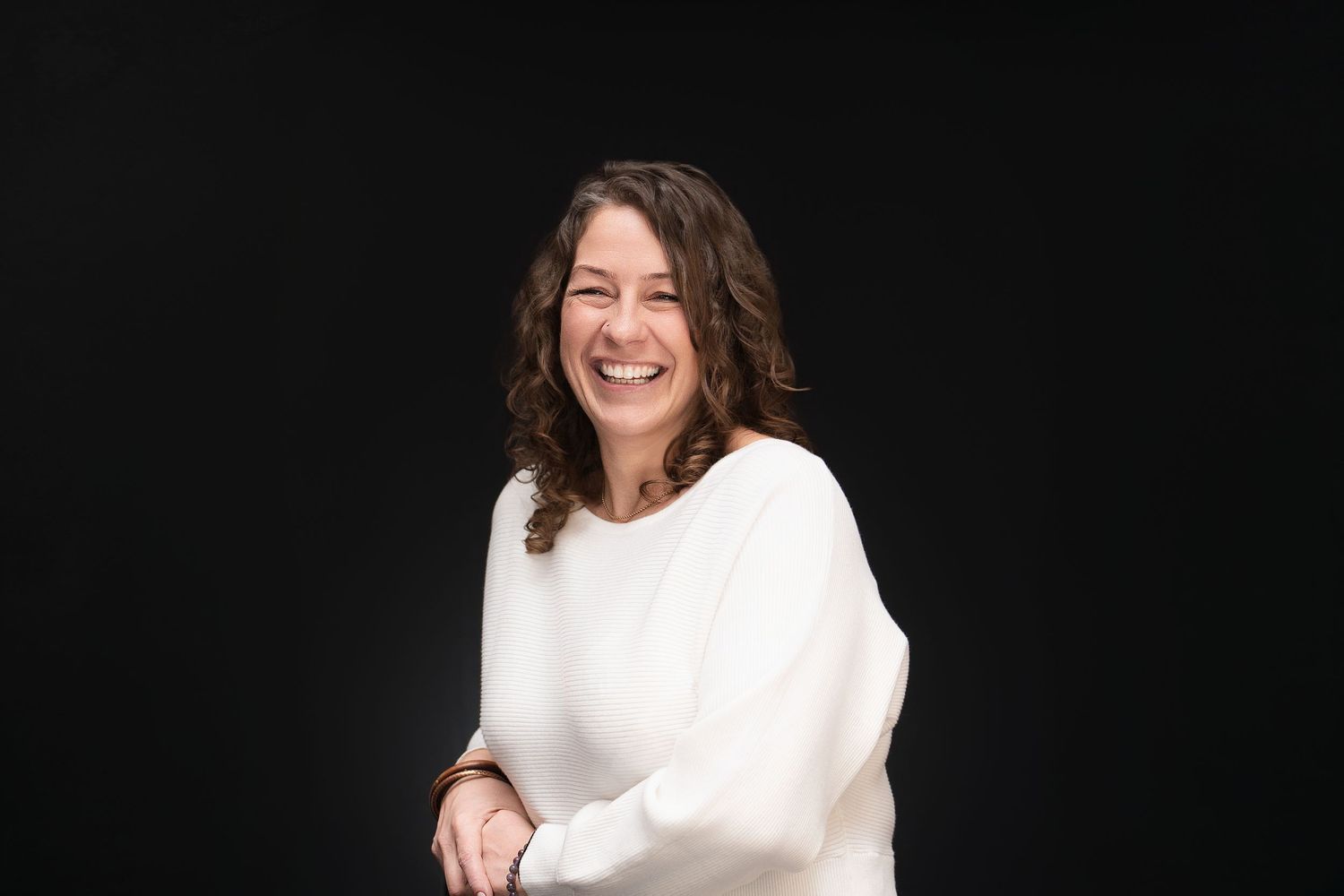 A smiling woman with dark hair wearing a white sweater poses confidently against a dark black background.
