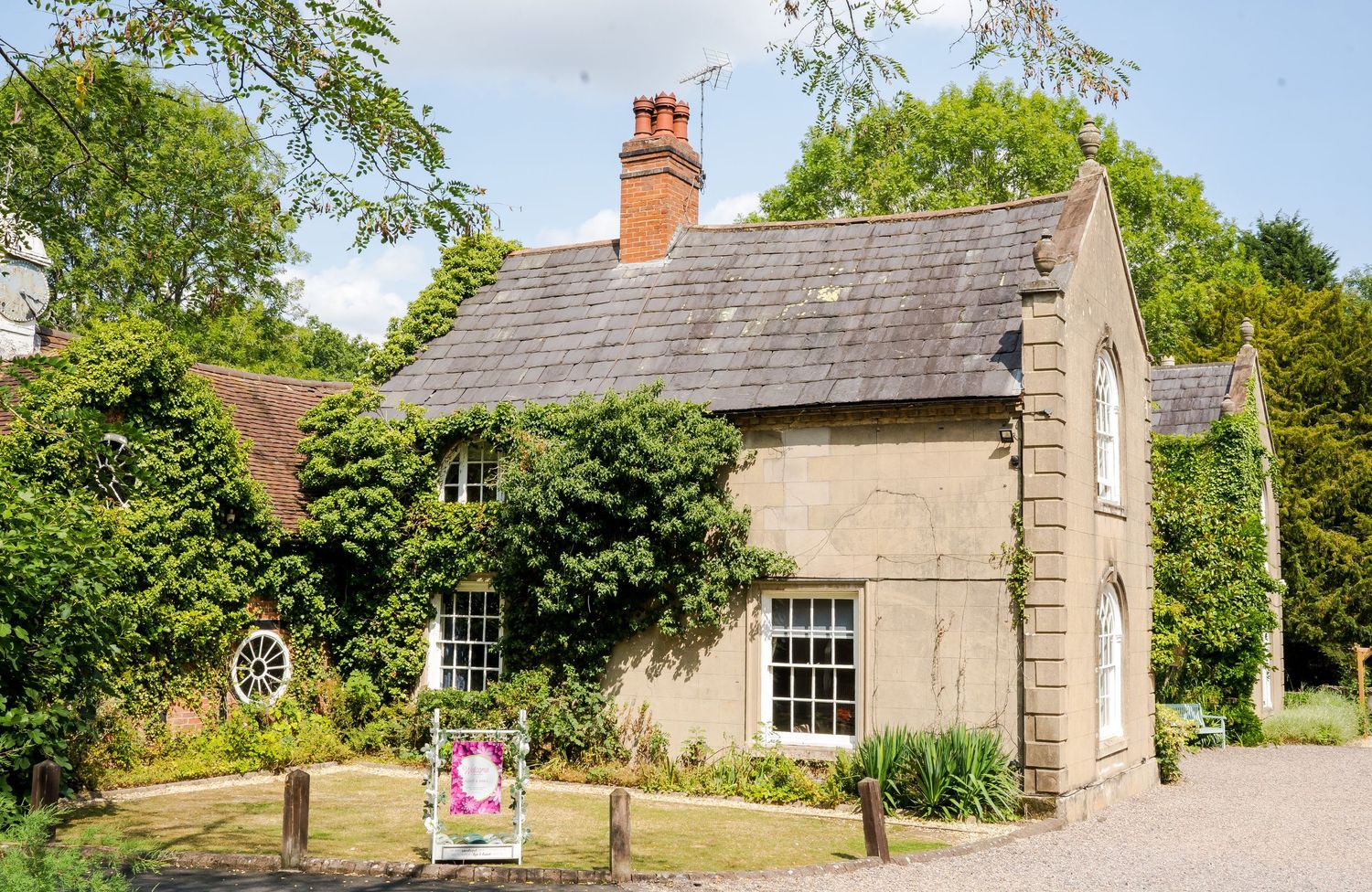 A charming stone cottage with ivy-covered walls, slate roof, and a colorful garden sign, surrounded by lush green trees.