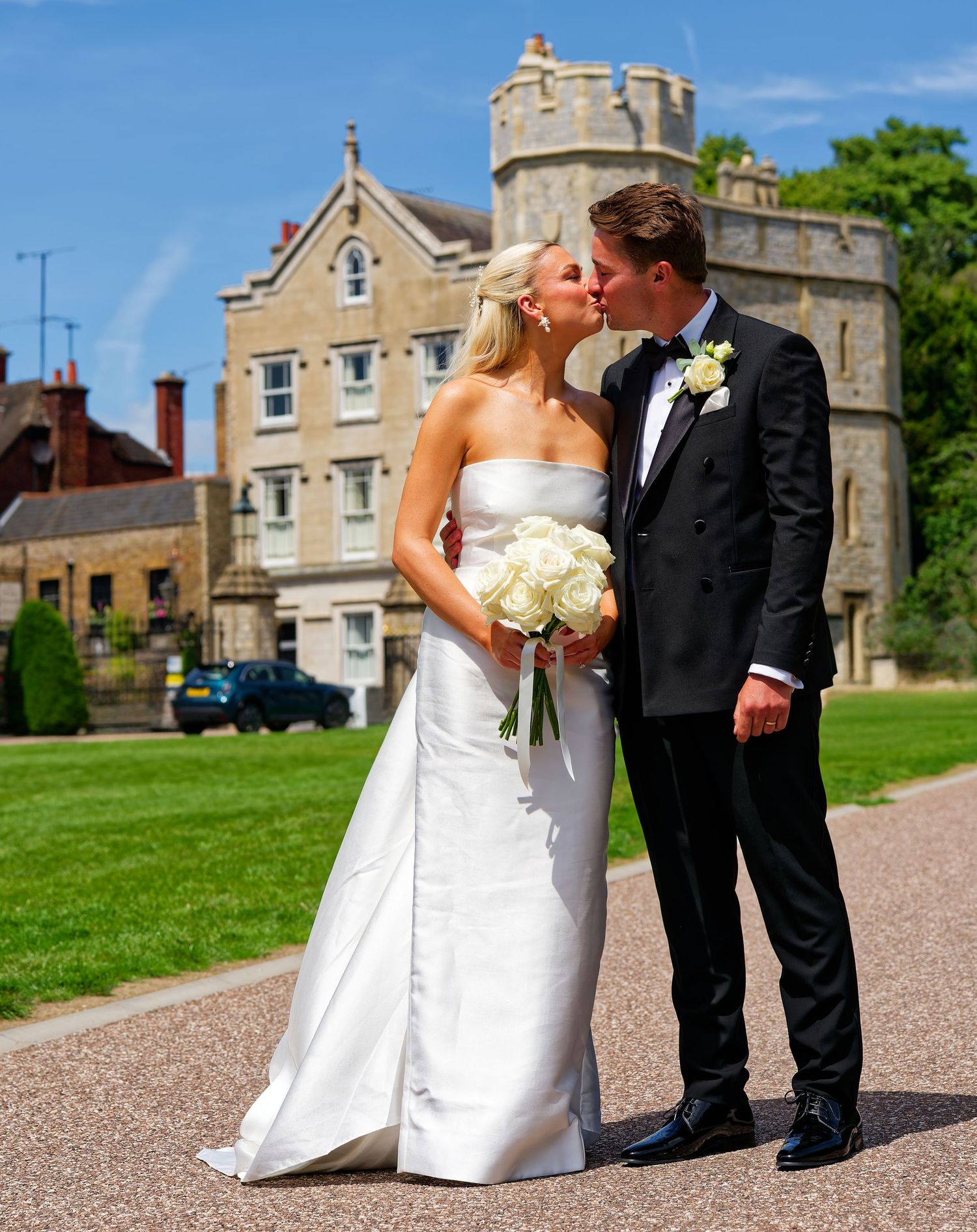 Bride and groom kissing in front of a historic stone castle on a sunny wedding day, bride holding white roses.