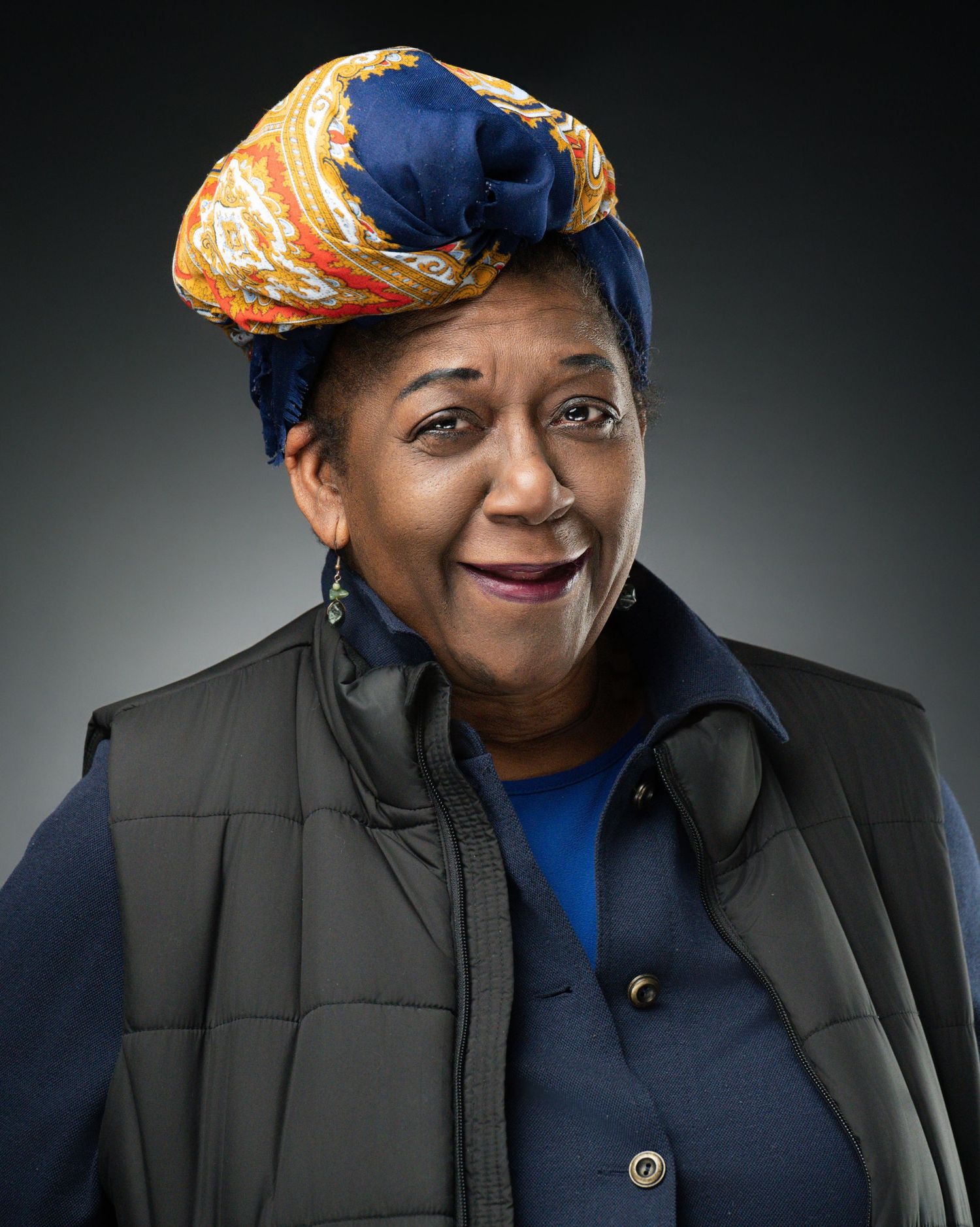 Smiling woman in blue headwrap and black quilted vest poses for portrait against dark background.