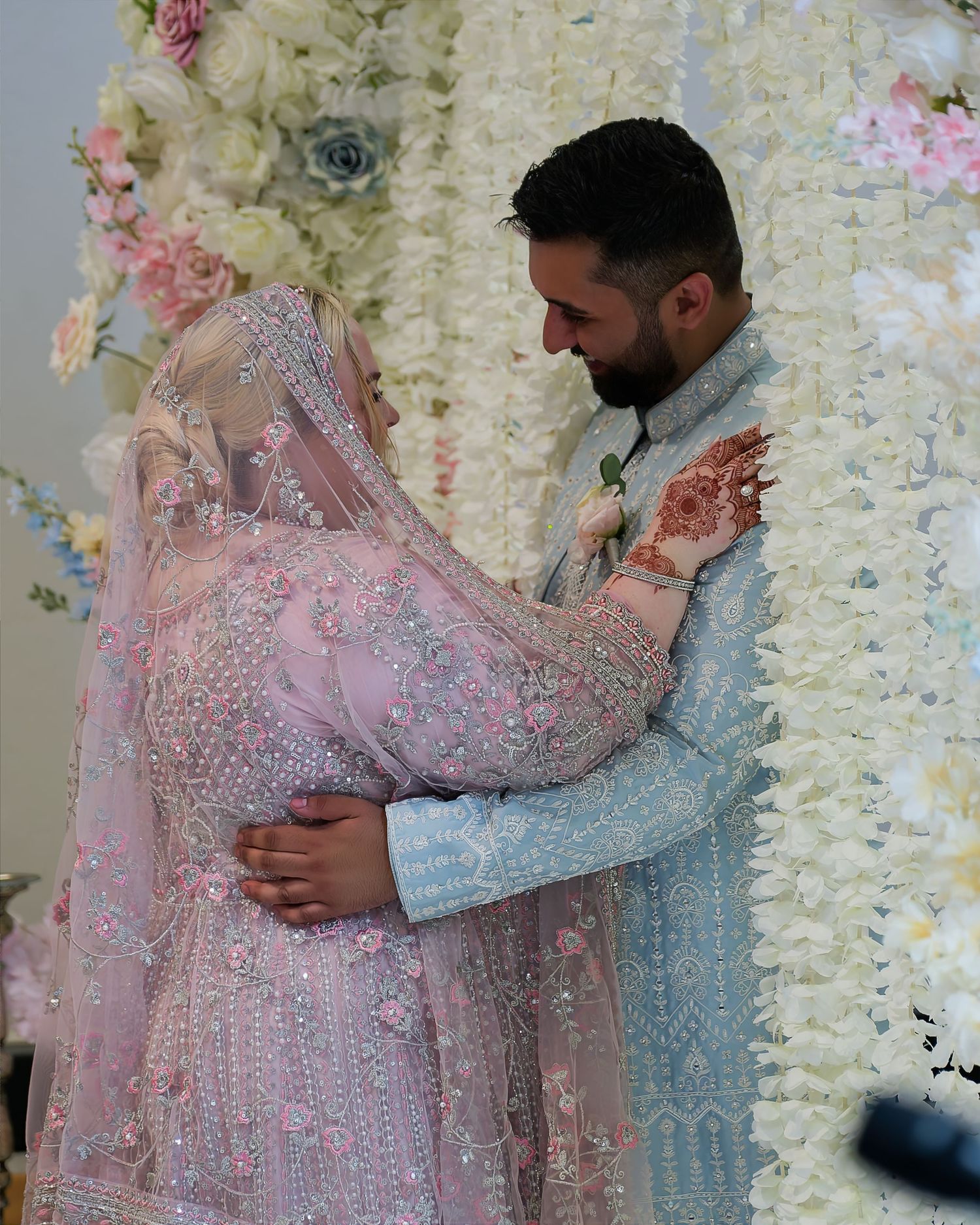 Couple in traditional South Asian wedding attire embrace tenderly under floral arch, bride in pink embroidered lehenga with veil.