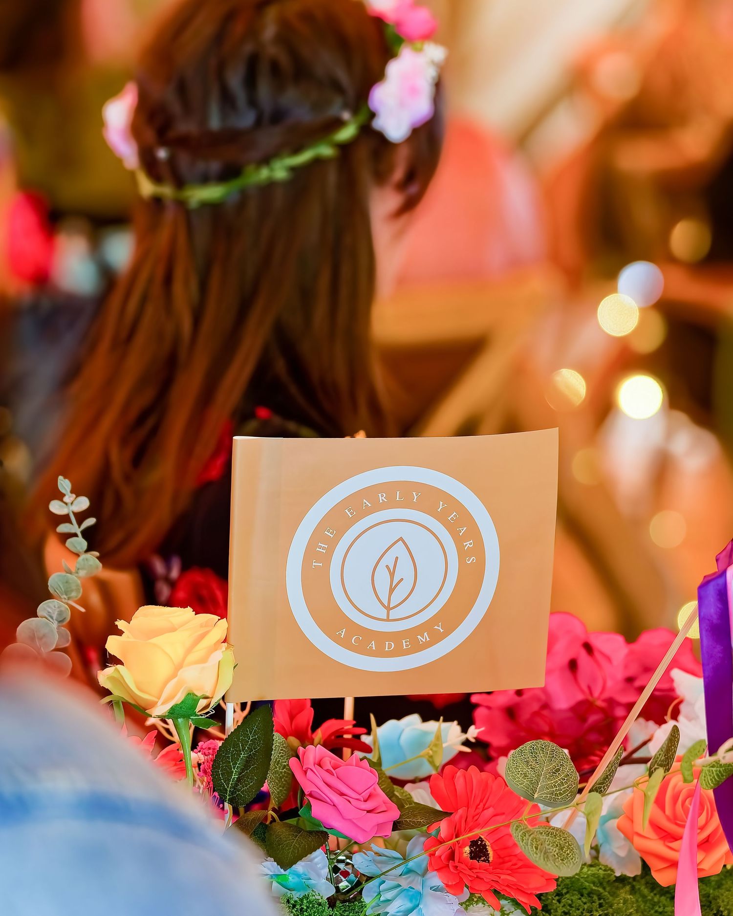 A floral arrangement with colorful roses surrounds a sign for The Laurel Tree Academy, with a woman wearing a flower crown behind it.