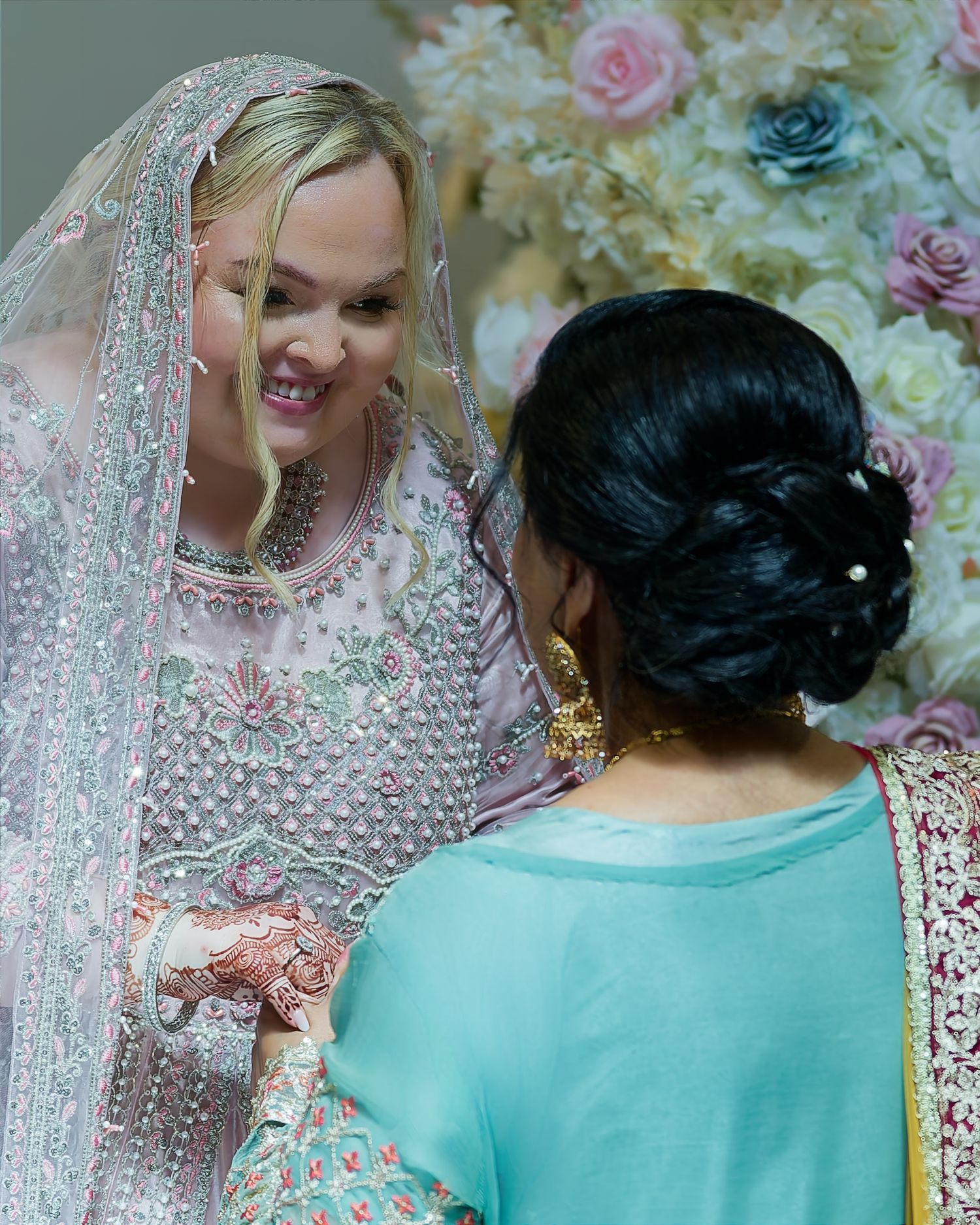 Bride in embroidered veil with mehndi hands smiles at guest in teal sari at floral Indian wedding ceremony.