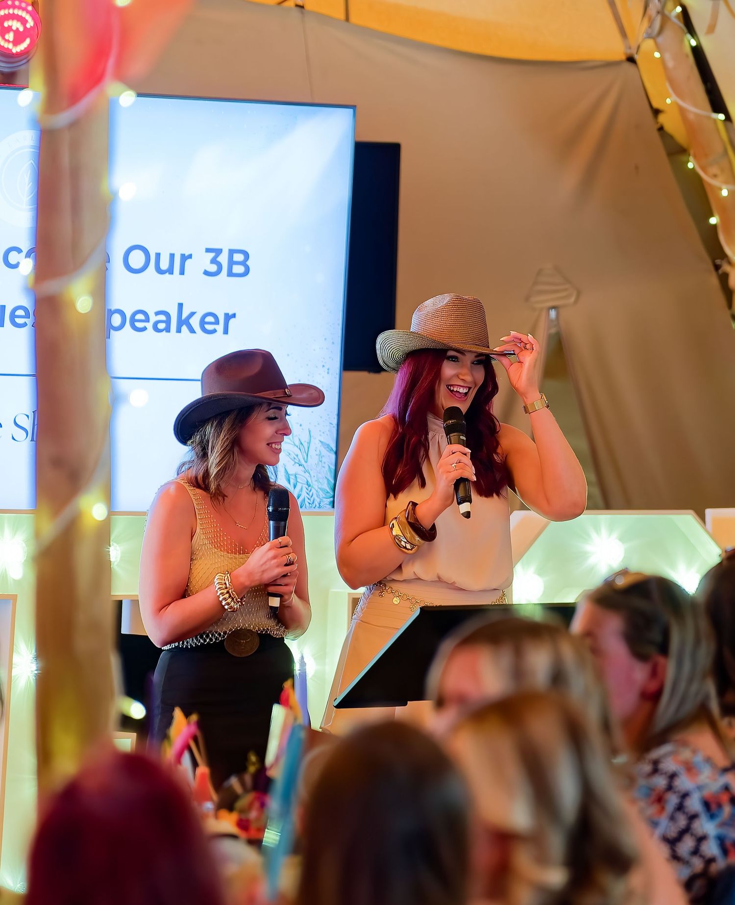 Two women wearing cowboy hats speak into microphones on stage at an indoor event under a tent.