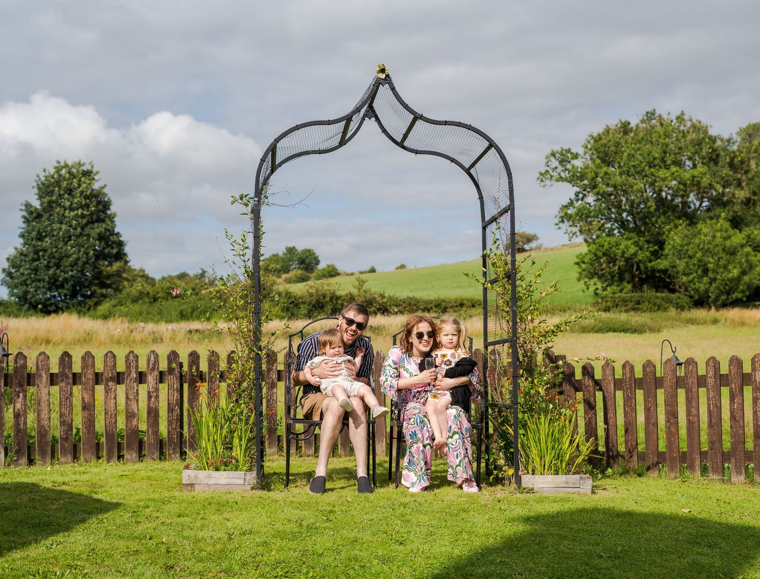 A decorative metal garden arch stands in a grassy backyard with a wooden picket fence and green hills in the background.