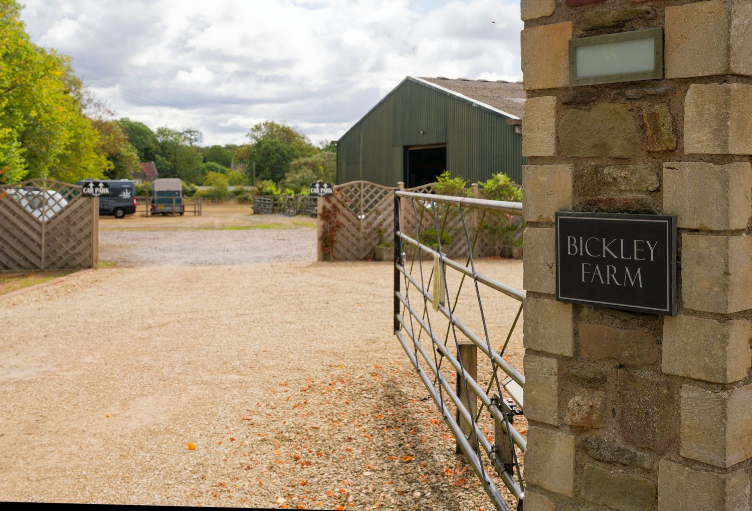 Entrance gate to Bickley Farm with stone pillar, metal gate, gravel driveway, and green barn in background.