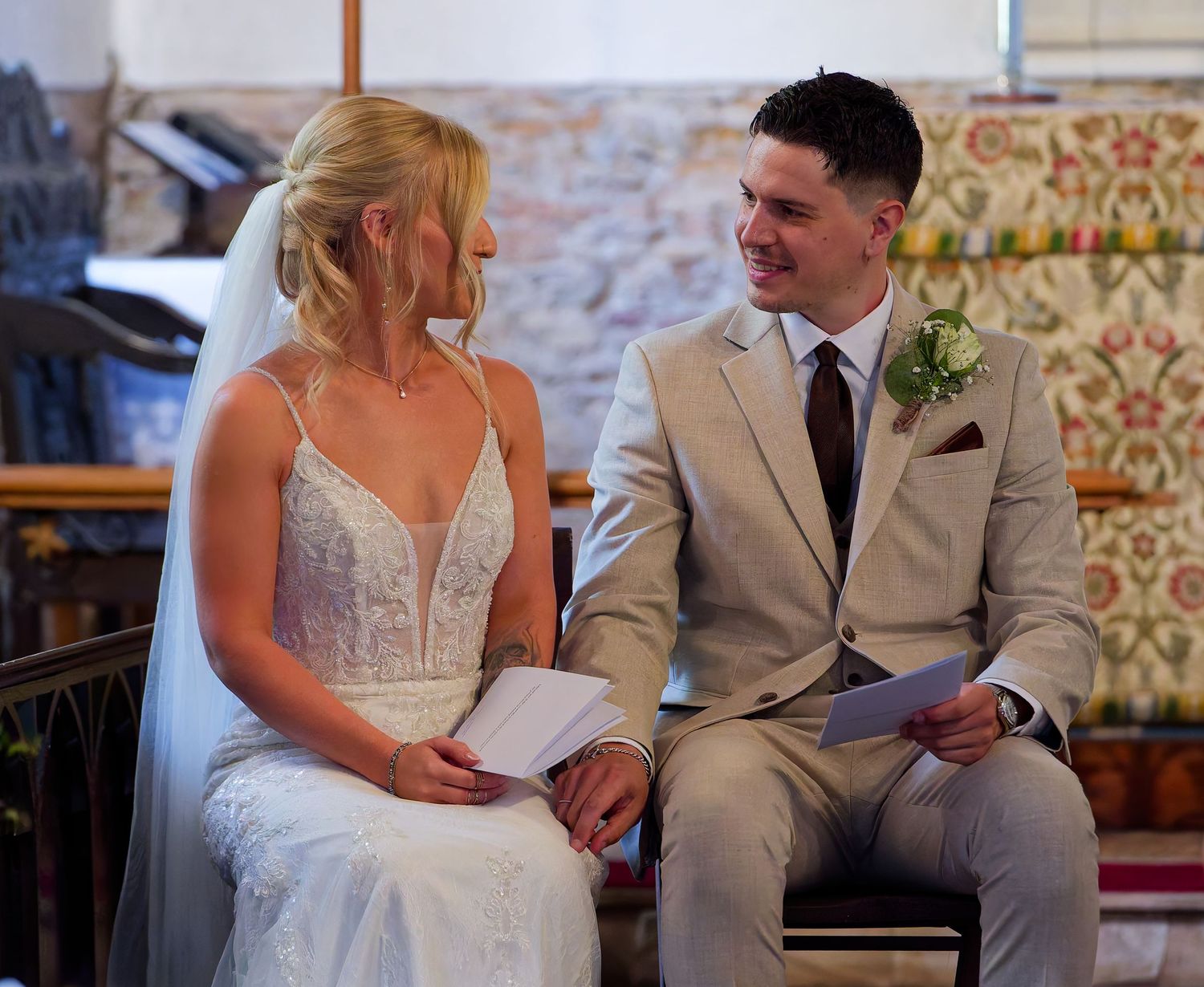 Bride and groom sitting together smiling during wedding ceremony, bride in white lace gown, groom in beige suit.