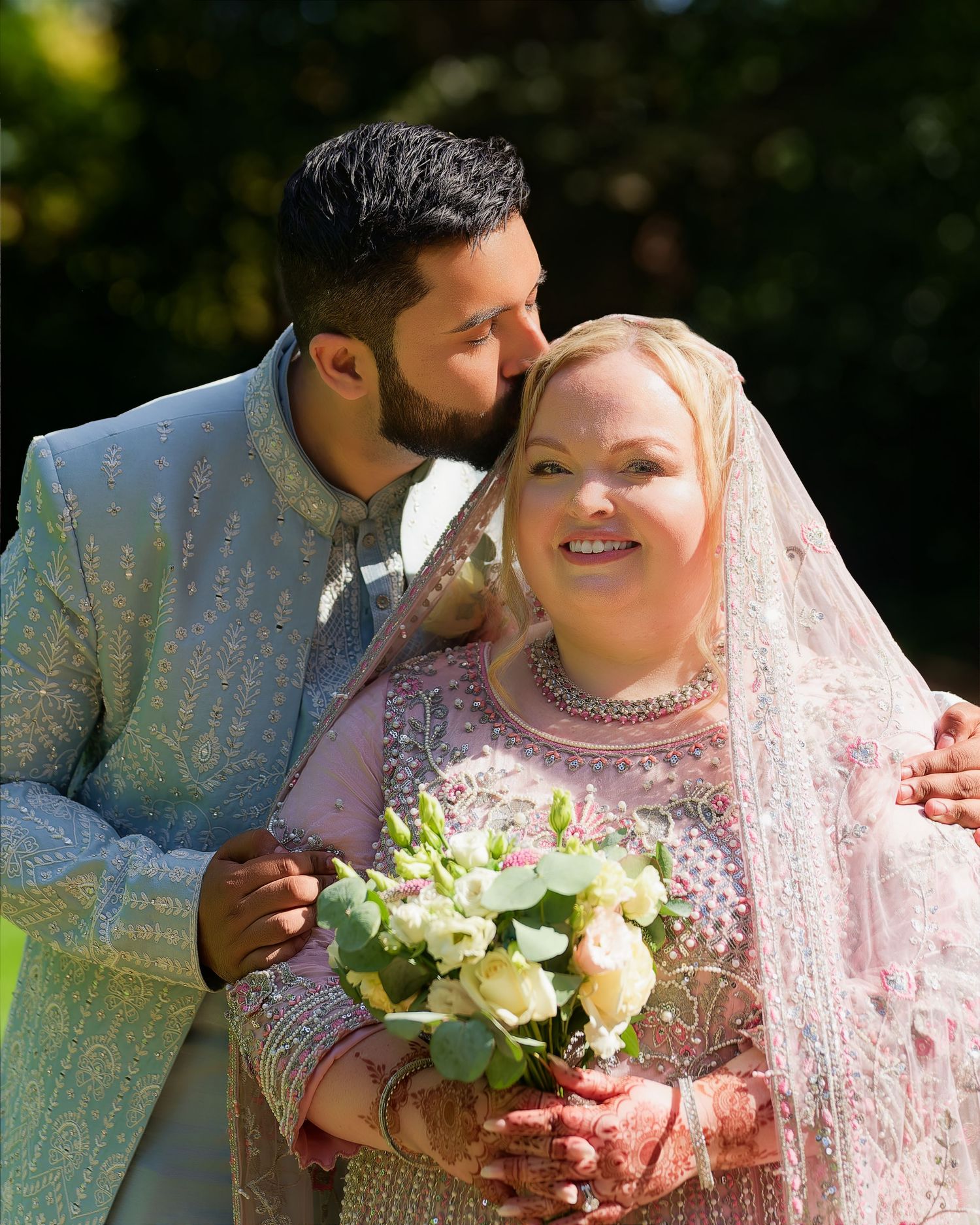 Groom in light blue sherwani kisses bride wearing pink embroidered lehenga holding white bouquet outdoors.