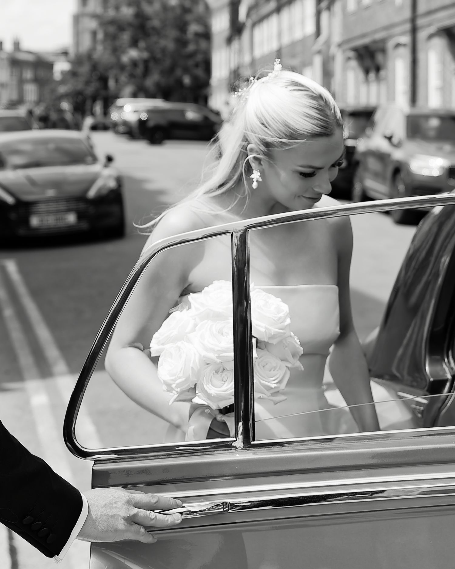 Black and white photo of a bride in a strapless gown holding white roses, stepping into a vintage car on a sunny day.