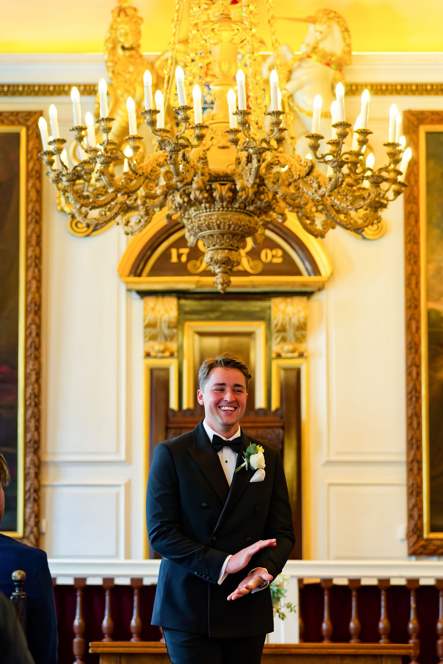 Smiling groom in tuxedo stands beneath ornate chandelier in elegant wedding venue.