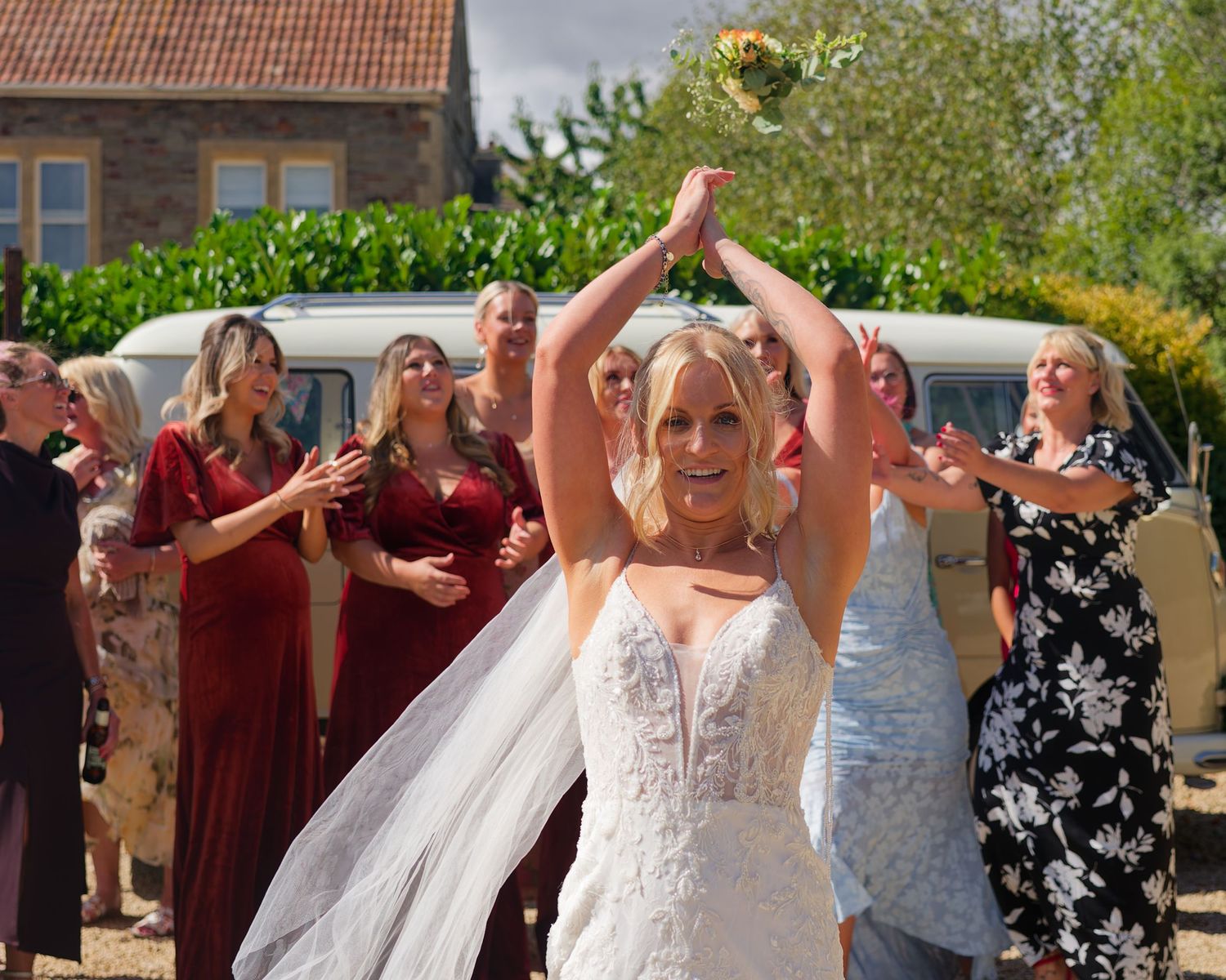 Bride in white lace gown tosses bouquet to guests in burgundy dresses at outdoor wedding celebration.