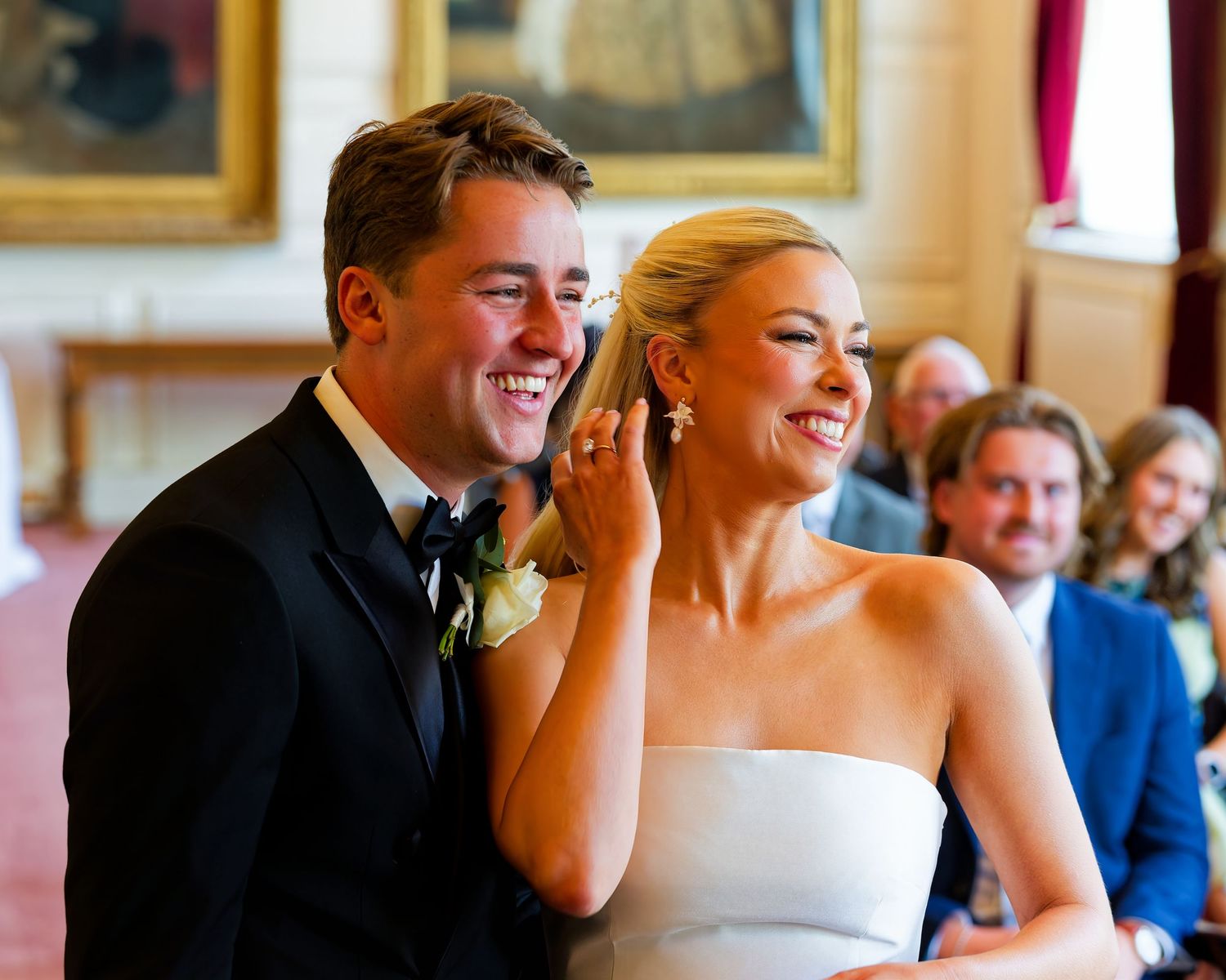 Happy couple laughing together during wedding ceremony, bride in strapless gown, groom in black tuxedo, elegant venue.