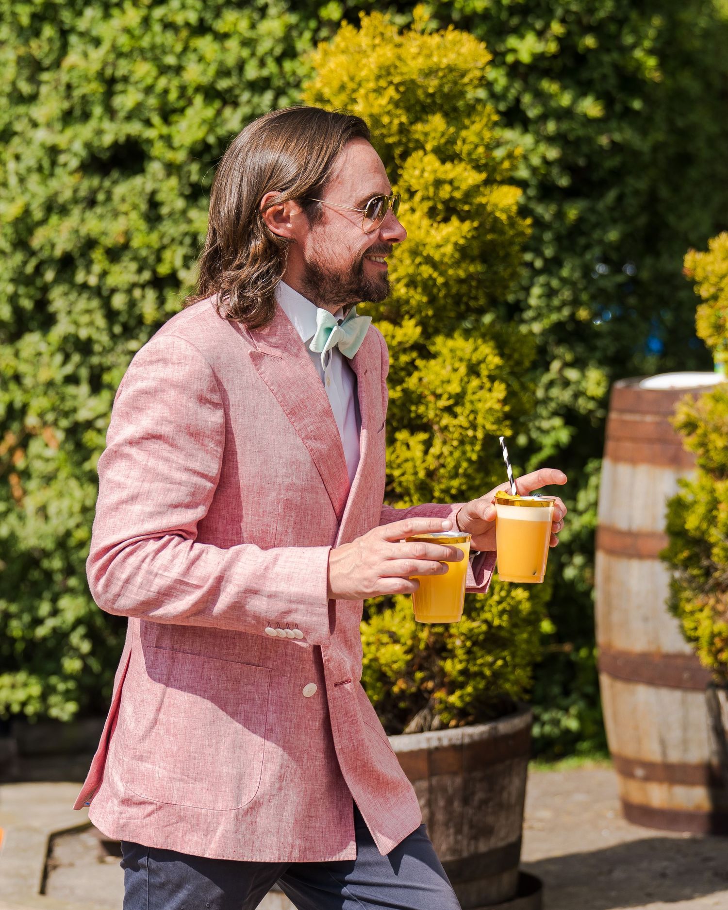 A person in a pink blazer and sunglasses holds an orange cocktail while standing outdoors near yellow flowers.