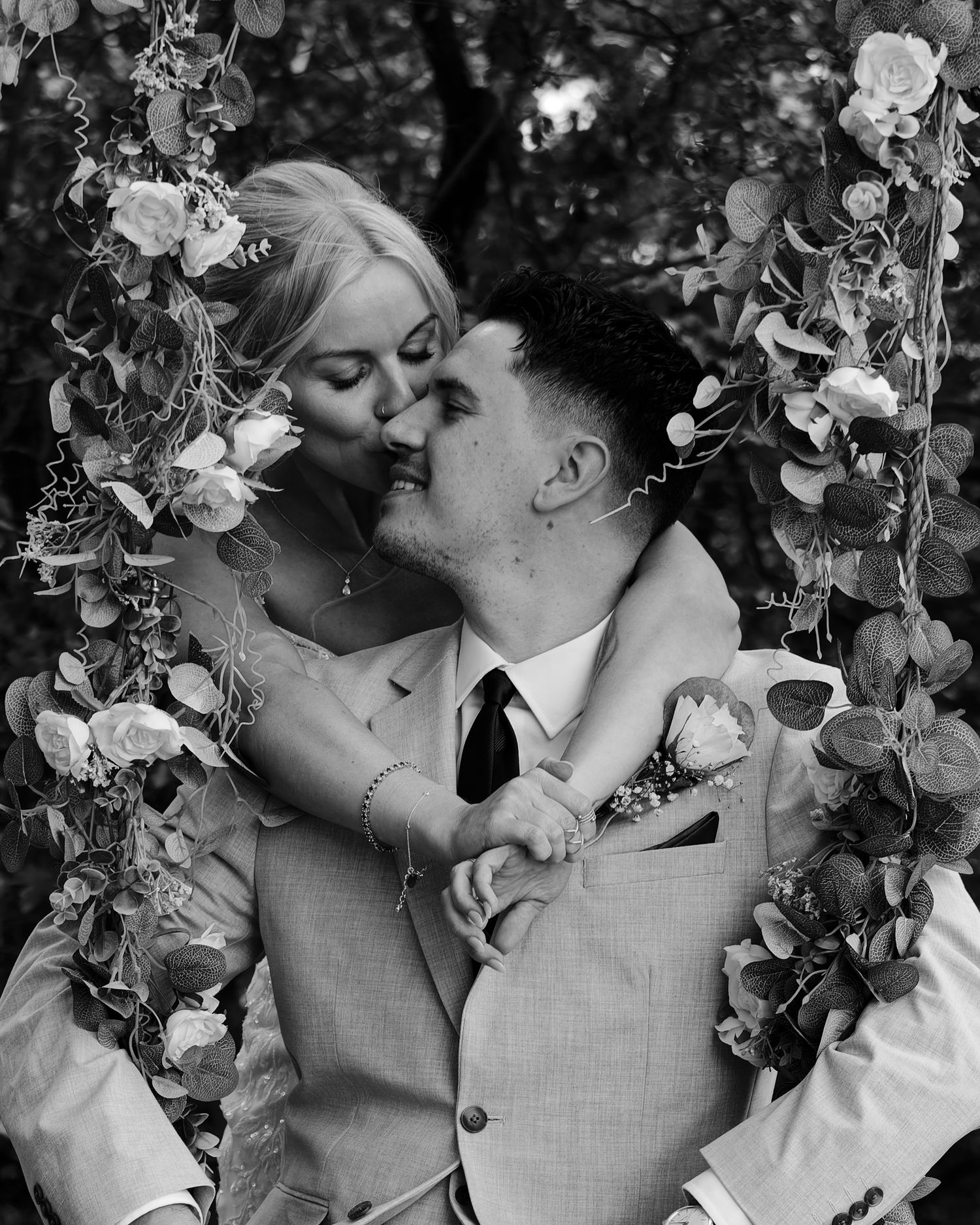 Black and white photo of a couple kissing on a floral swing decorated with roses and greenery at their wedding.