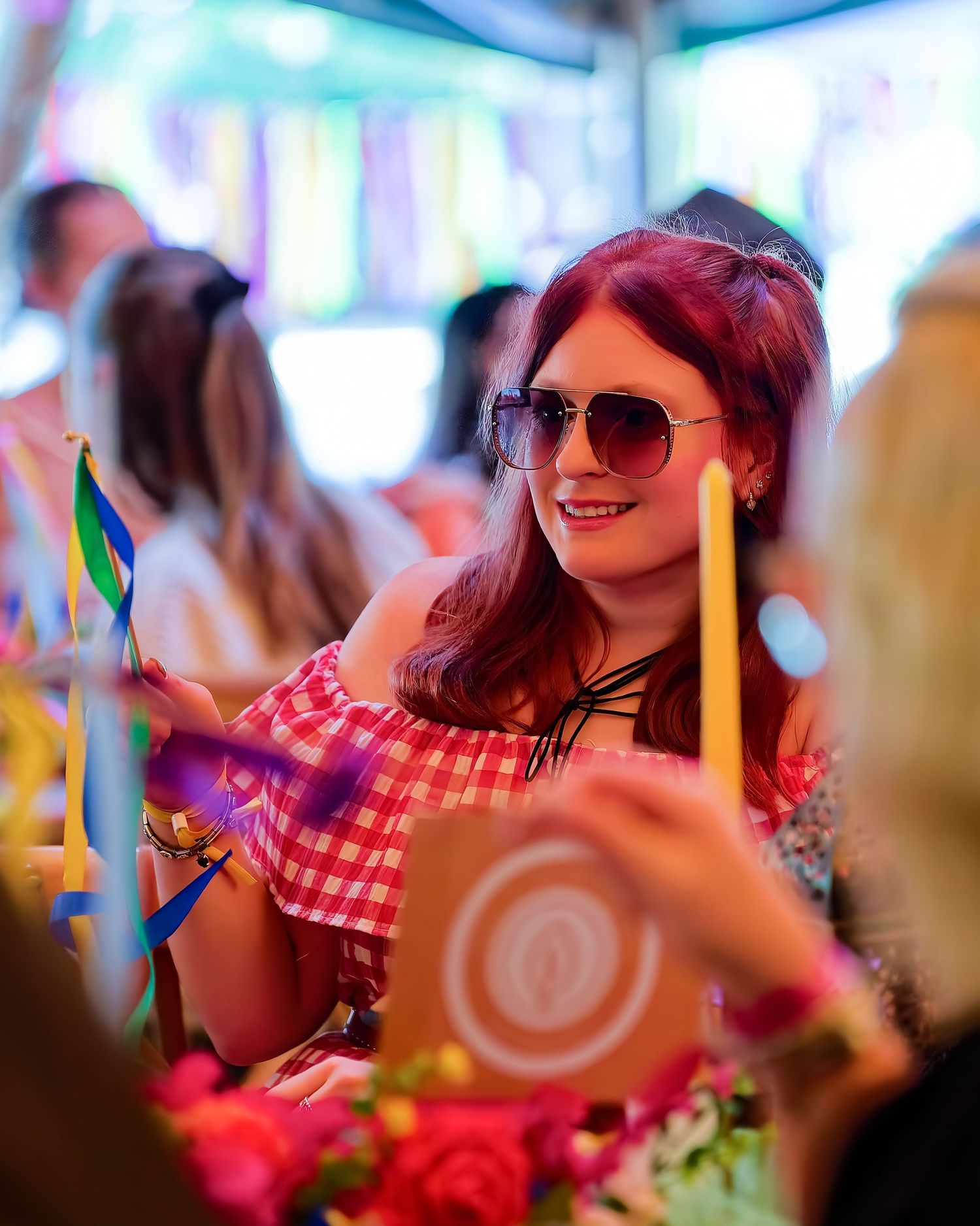 A woman with red hair and aviator sunglasses smiles at a colorful festival event, wearing a red gingham off-shoulder top.
