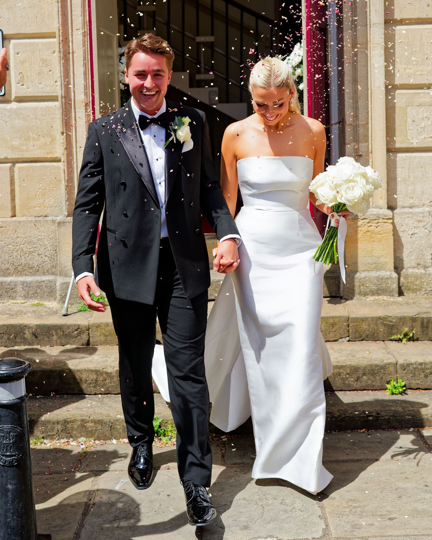 Newlywed couple exits church in confetti shower, groom in black tuxedo, bride in white strapless gown holding white bouquet.