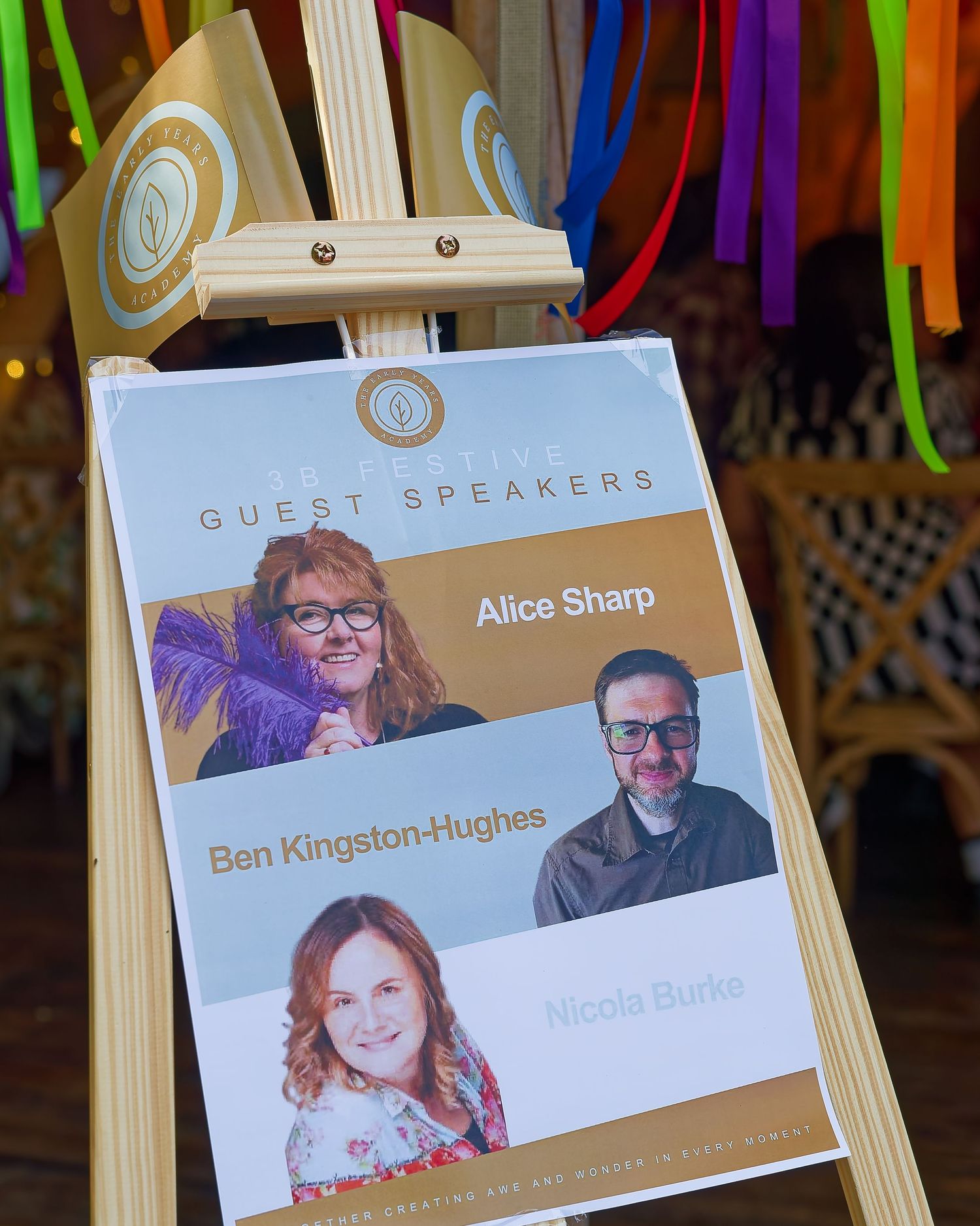 A guest speakers board on an easel featuring Alice Sharp, Ben Kingston-Hughes, and Nicola Burke at a colorful event.