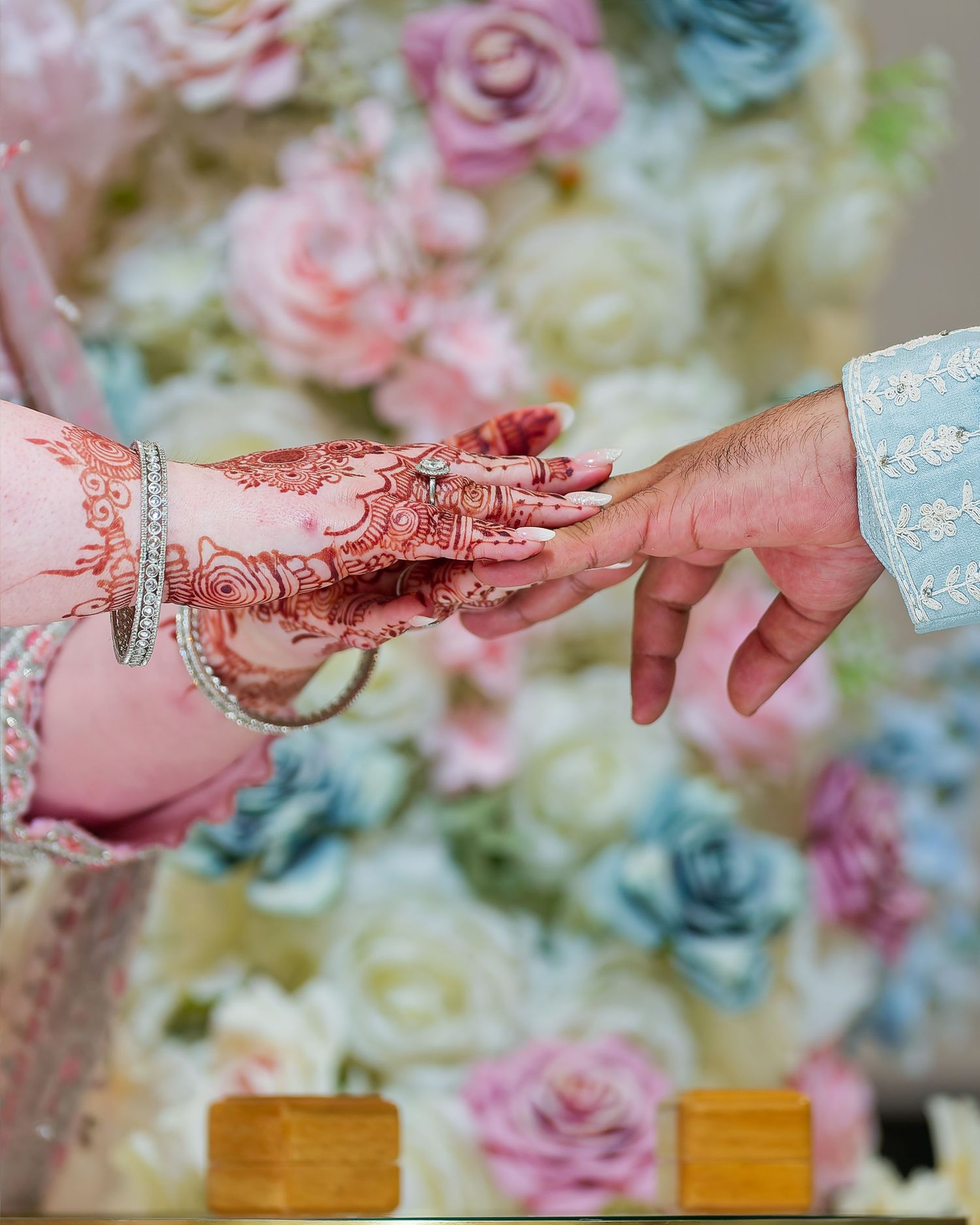 Bride with intricate henna mehndi design and bangles exchanging rings with groom against floral backdrop.