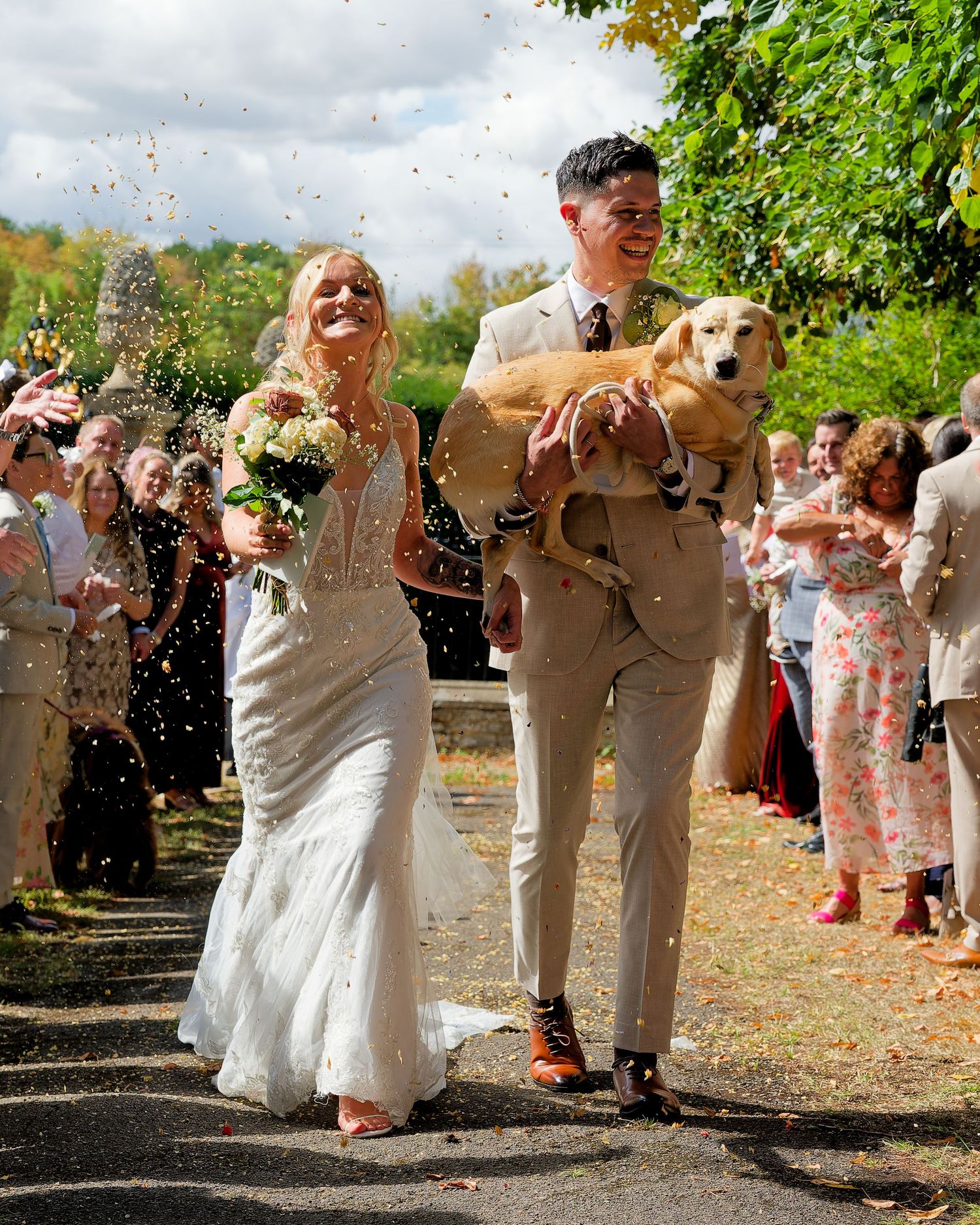 Joyful newlyweds walk through confetti with golden retriever as guests celebrate outdoor autumn wedding.