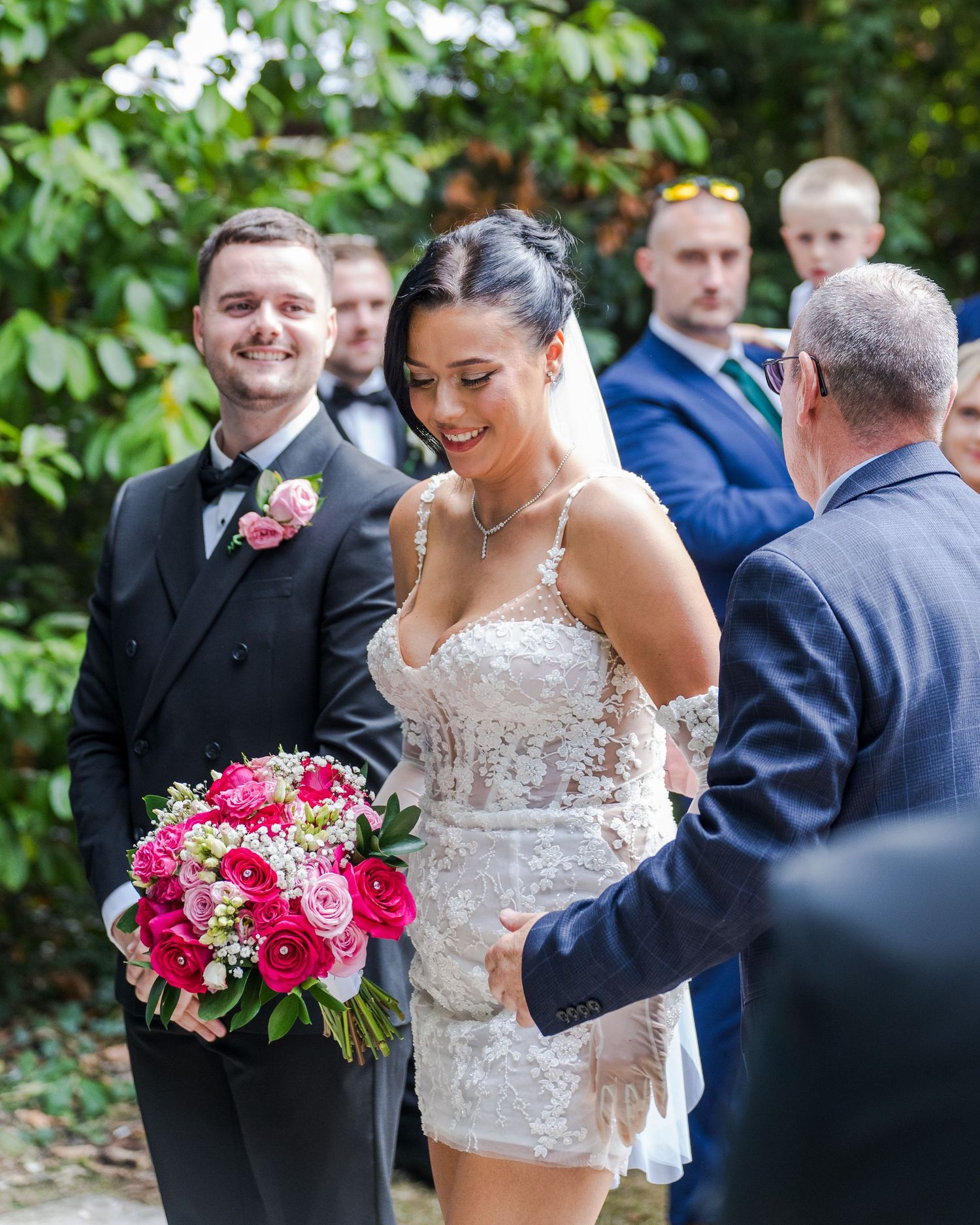 Bride in lace mini dress holding pink bouquet smiles during outdoor wedding ceremony with groom in black tuxedo nearby.