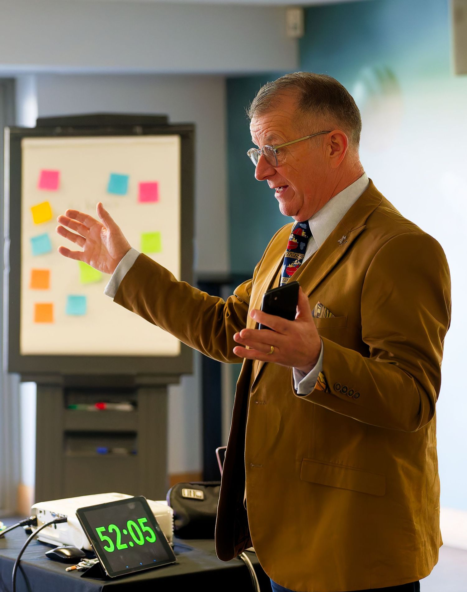 Presenter in a tan blazer and striped tie gestures while speaking beside a whiteboard of colorful sticky notes and a 52:05 countdown timer.