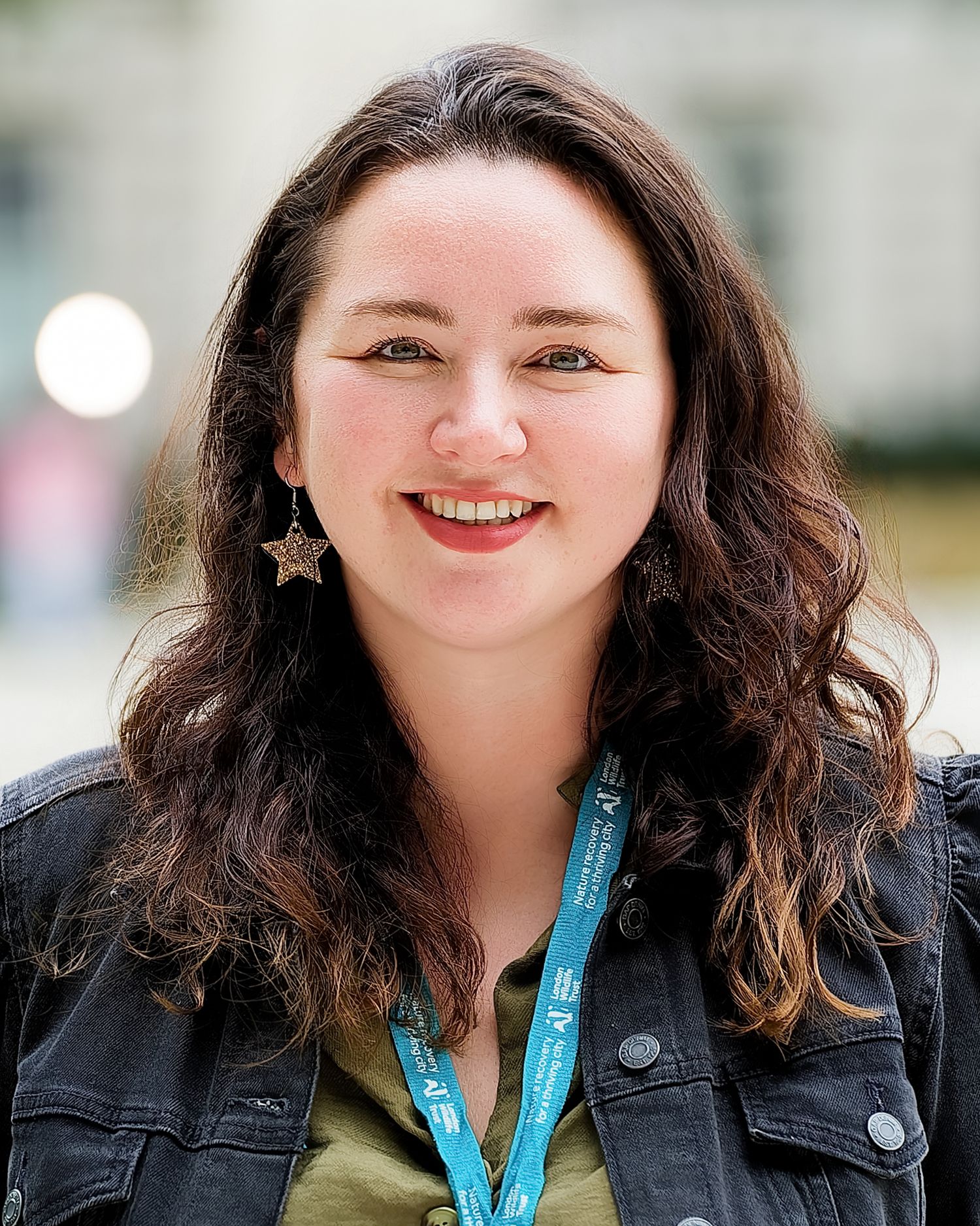 Smiling young woman with curly brown hair, star earrings, denim jacket, and teal lanyard outdoors.