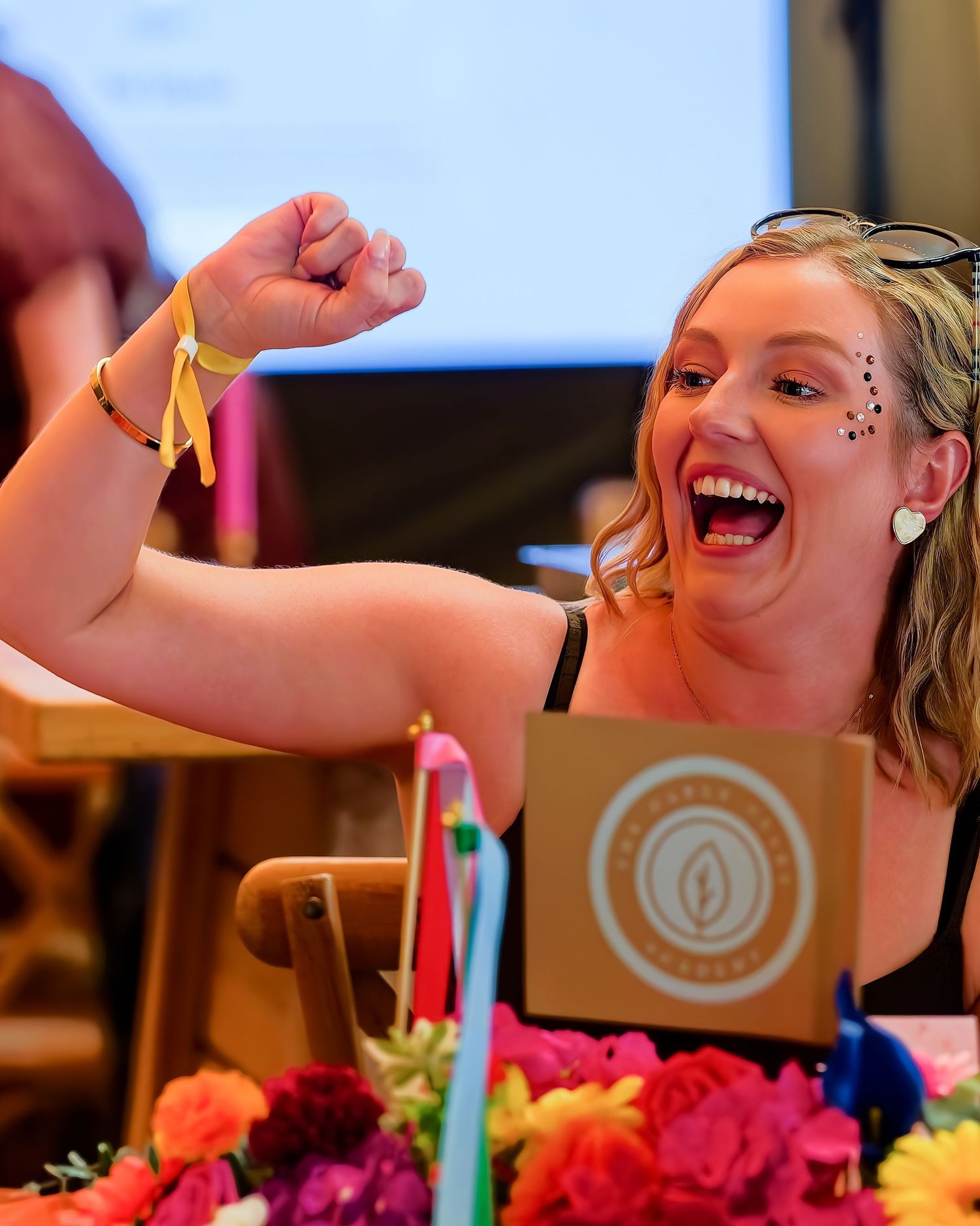 Excited woman flexing her arm and laughing at a colorful event table with flowers and a branded sign.