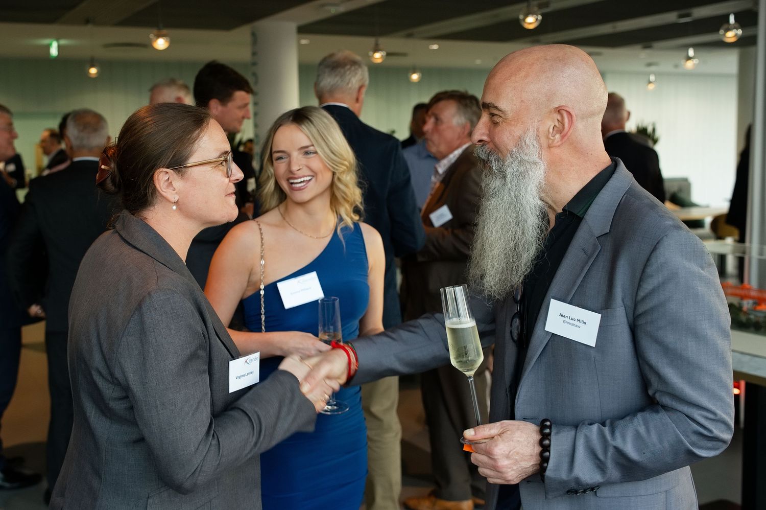 Bald bearded man holding a champagne glass shakes hands with a smiling woman in glasses at a crowded networking reception.