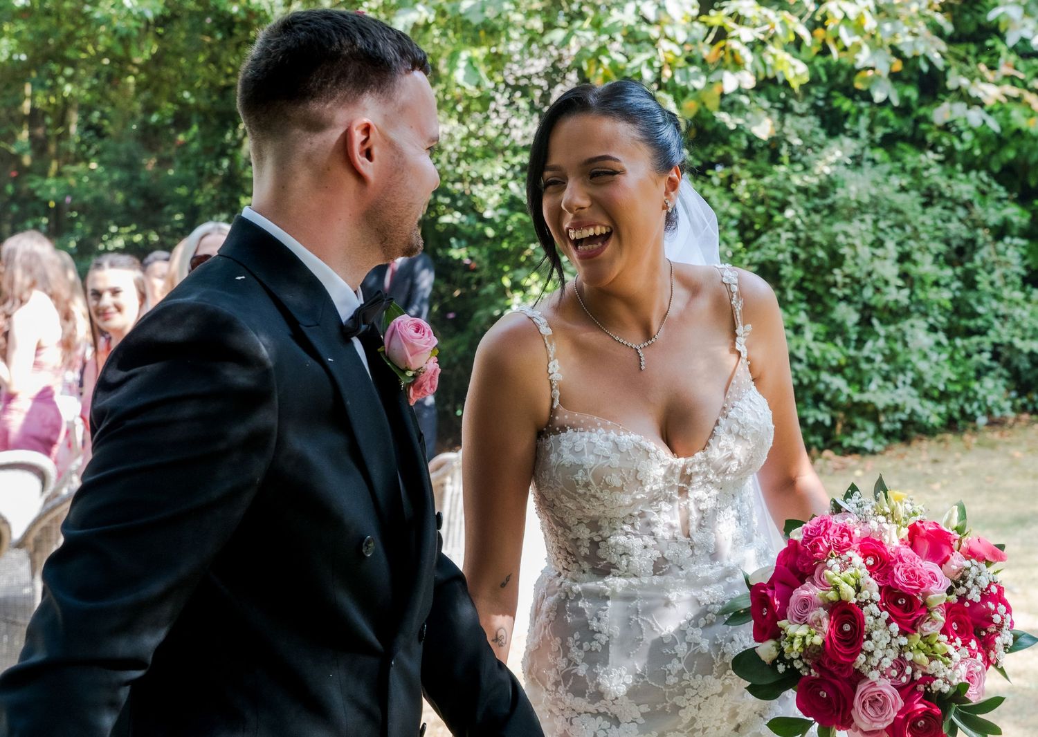 Bride in lace gown holding pink bouquet laughs with groom in black tuxedo at outdoor wedding ceremony.