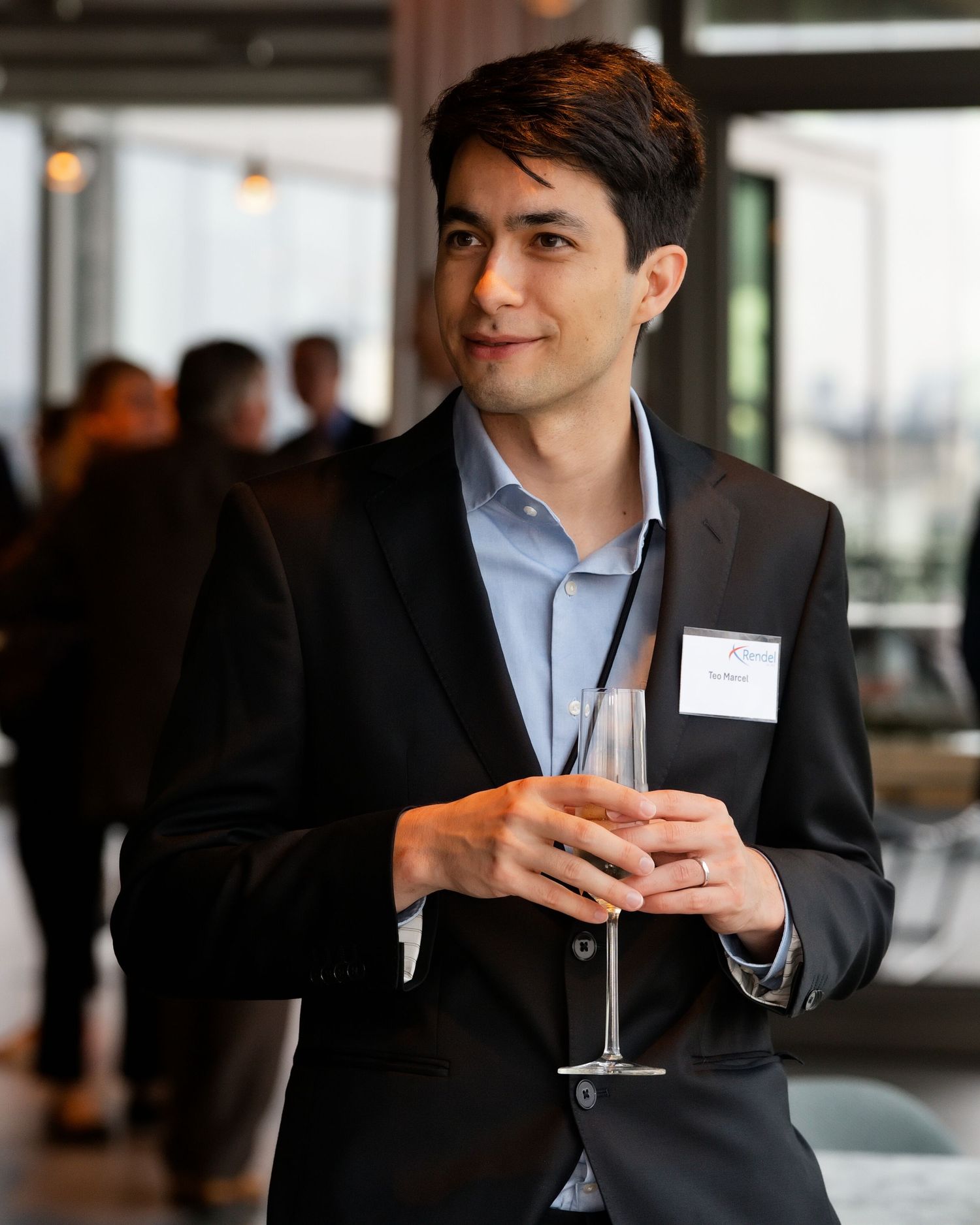 Man in a dark blazer and light blue shirt holds a champagne flute at a networking event with a name tag and guests behind him.