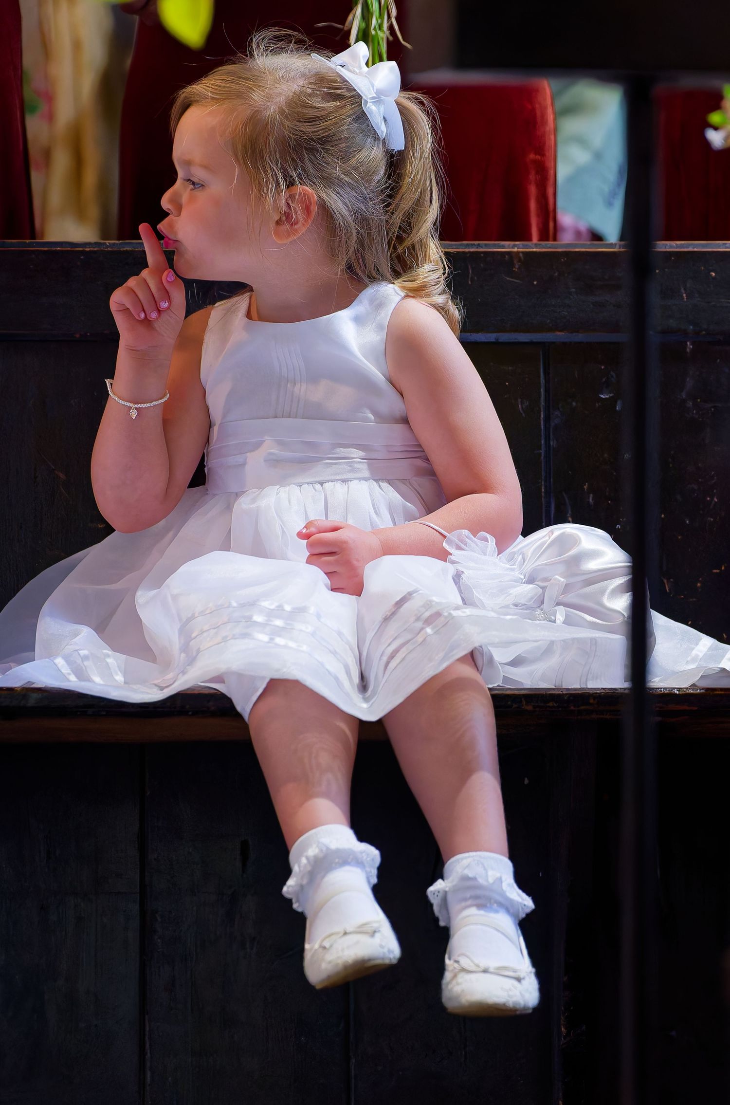Adorable flower girl in white dress and bow sits on a wooden bench with finger on her lips.