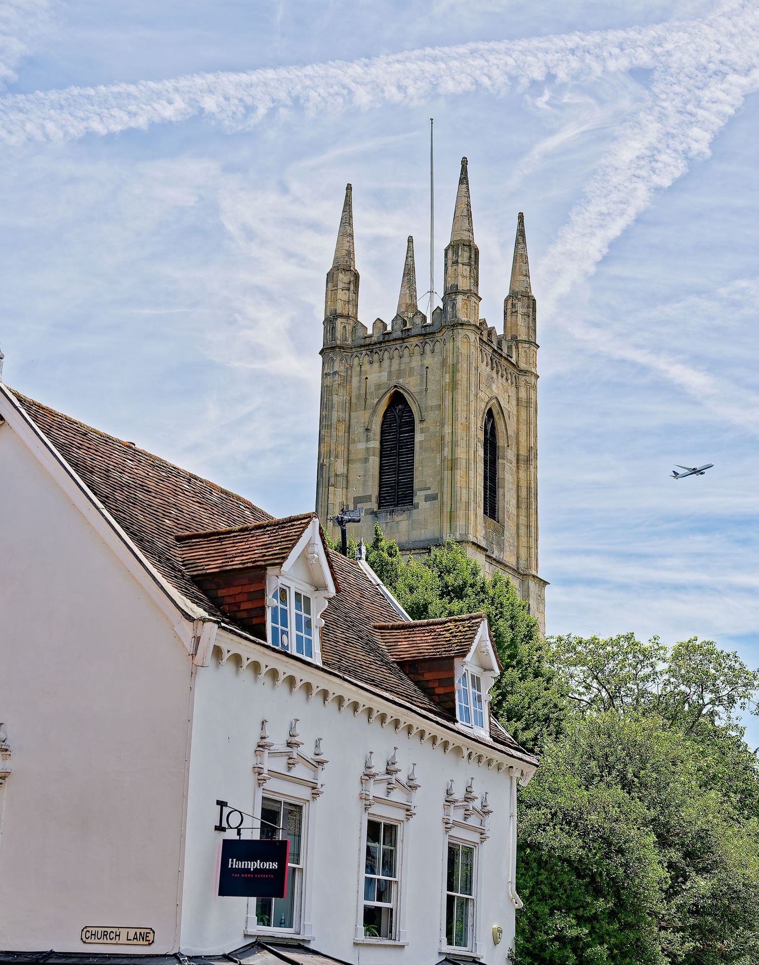 Historic Gothic church tower rises above white Georgian buildings on Church Lane, with a plane visible in the blue sky.