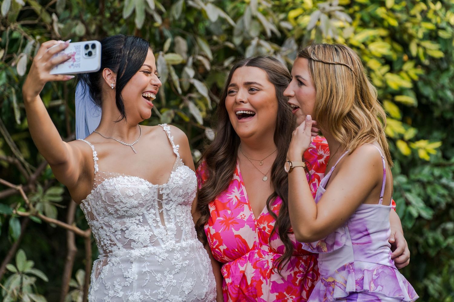 Bride in white lace gown takes selfie with two smiling bridesmaids in pink and purple dresses outdoors.
