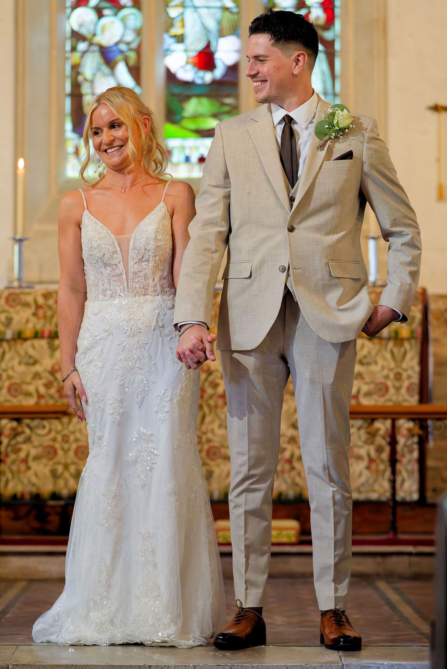 A bride in a lace gown and groom in a tan suit hold hands at the altar of a church.